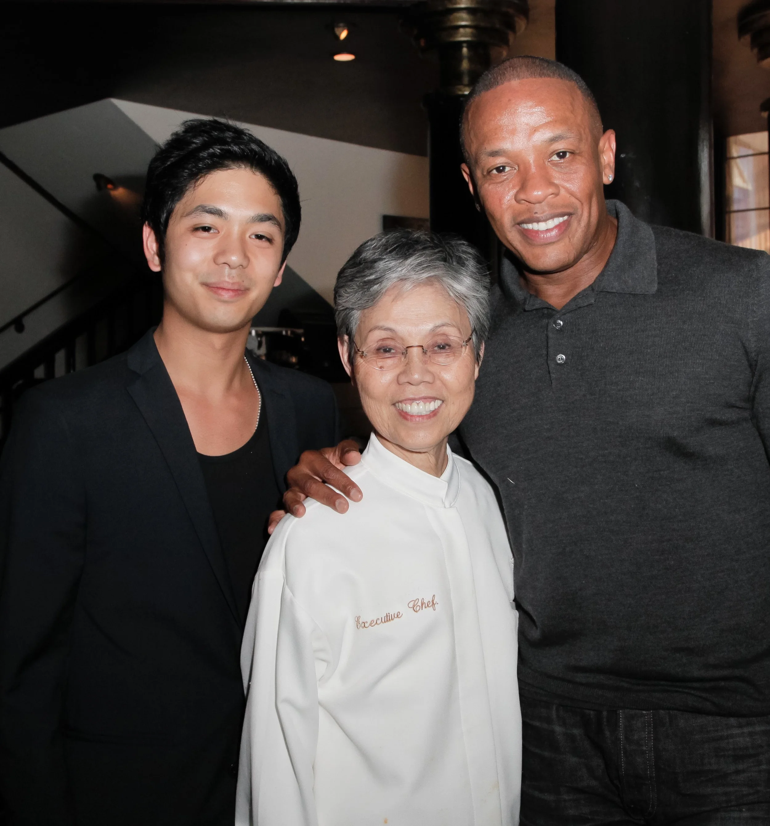 Three people smiling, a young man, an older woman in a chef's coat, and a tall man in a black polo shirt, posing together indoors.