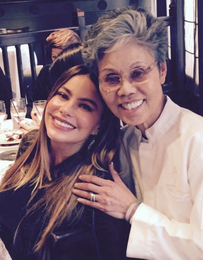 Two women smiling and posing for a photo at a restaurant, with tables set with glasses and food in the background.