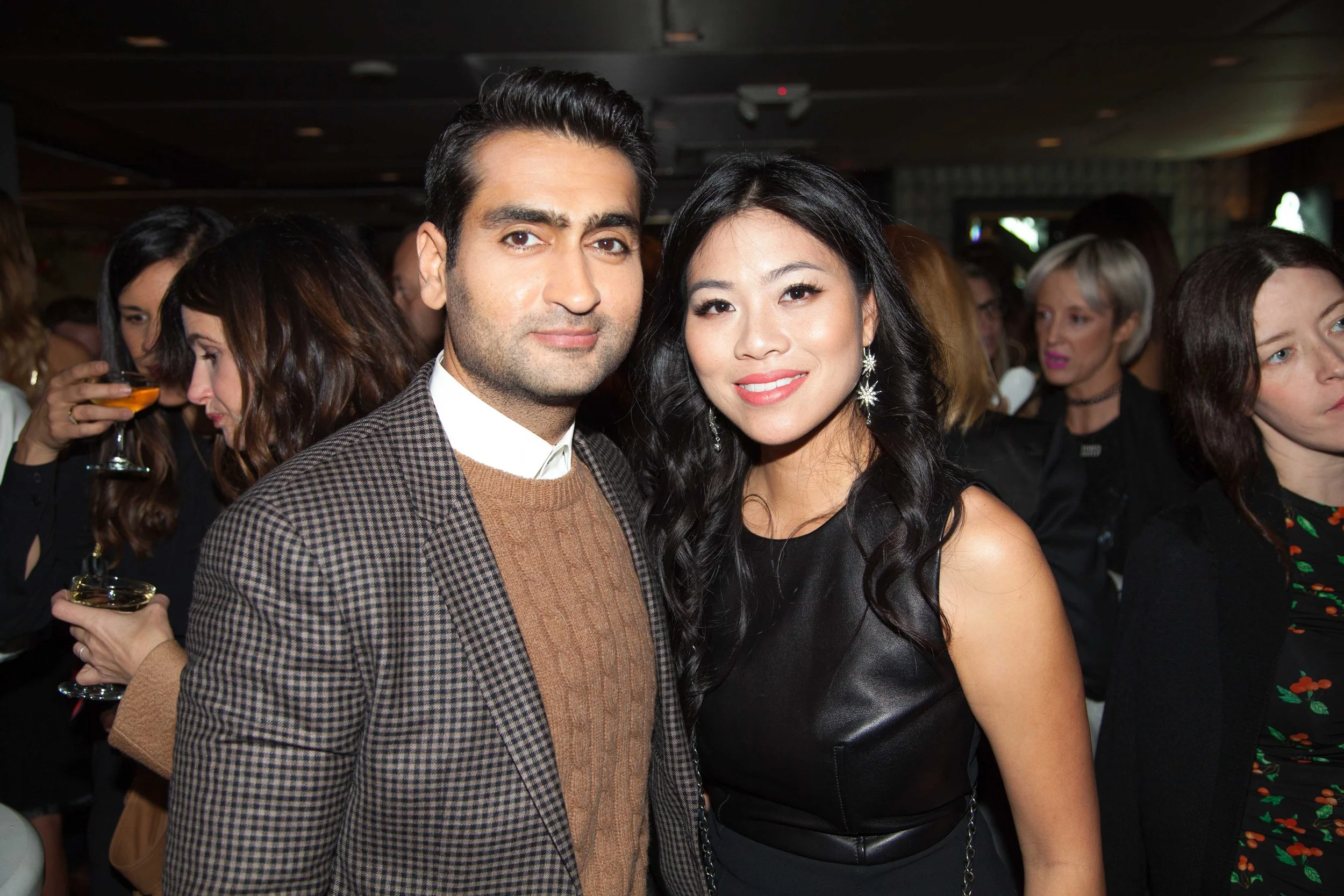 A man and a woman standing close together at a social event, surrounded by other people, some holding drinks, in an indoor setting.