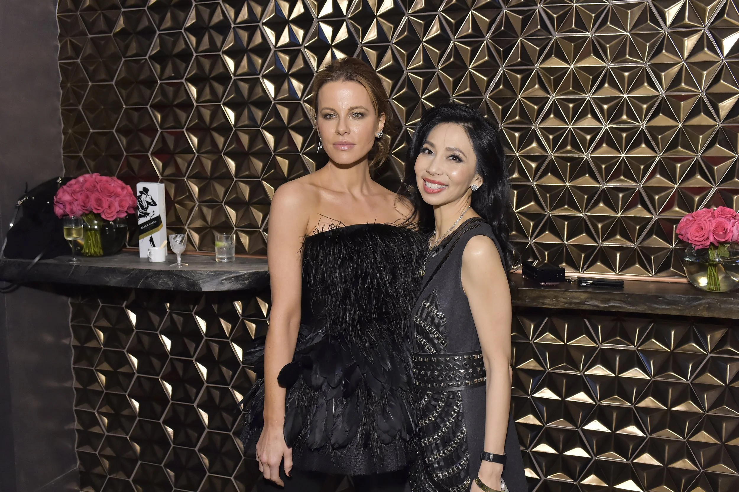 Two women in elegant black dresses posing at a stylish event with geometric gold wall decor, pink flower arrangements, and drinks on a marble counter.
