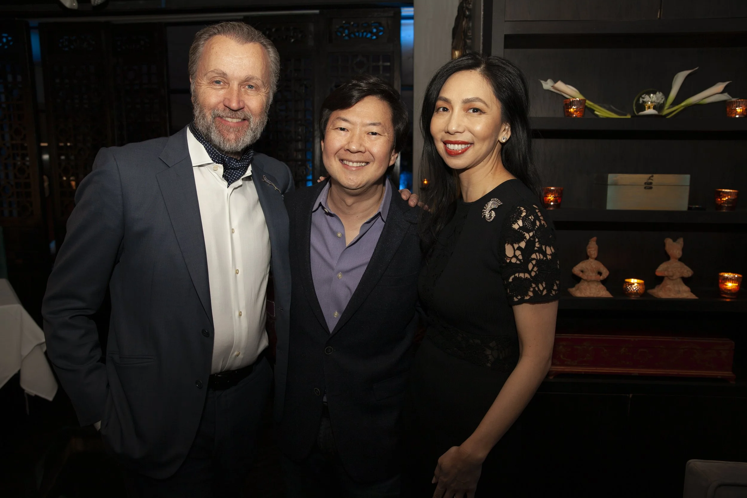 Three people smiling at a formal event, a man with gray hair and beard in a suit, a man with dark hair in a blazer, and a woman with long dark hair in a black dress with lace sleeves, standing close together in front of a dark background with shelves