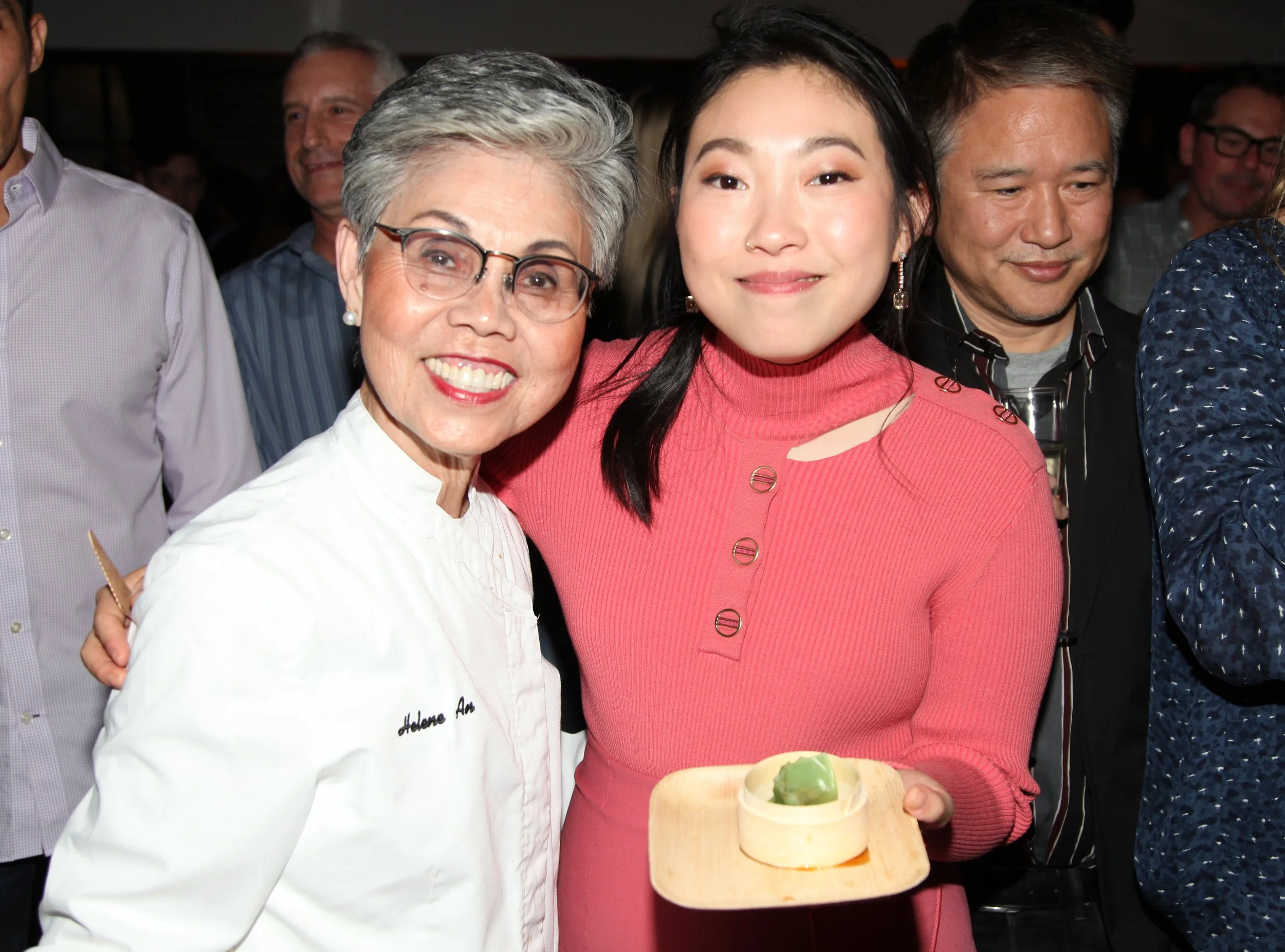 Two women smiling, one in a white chef's coat and the other in a pink sweater, holding a small dish with a green dessert at a social gathering.