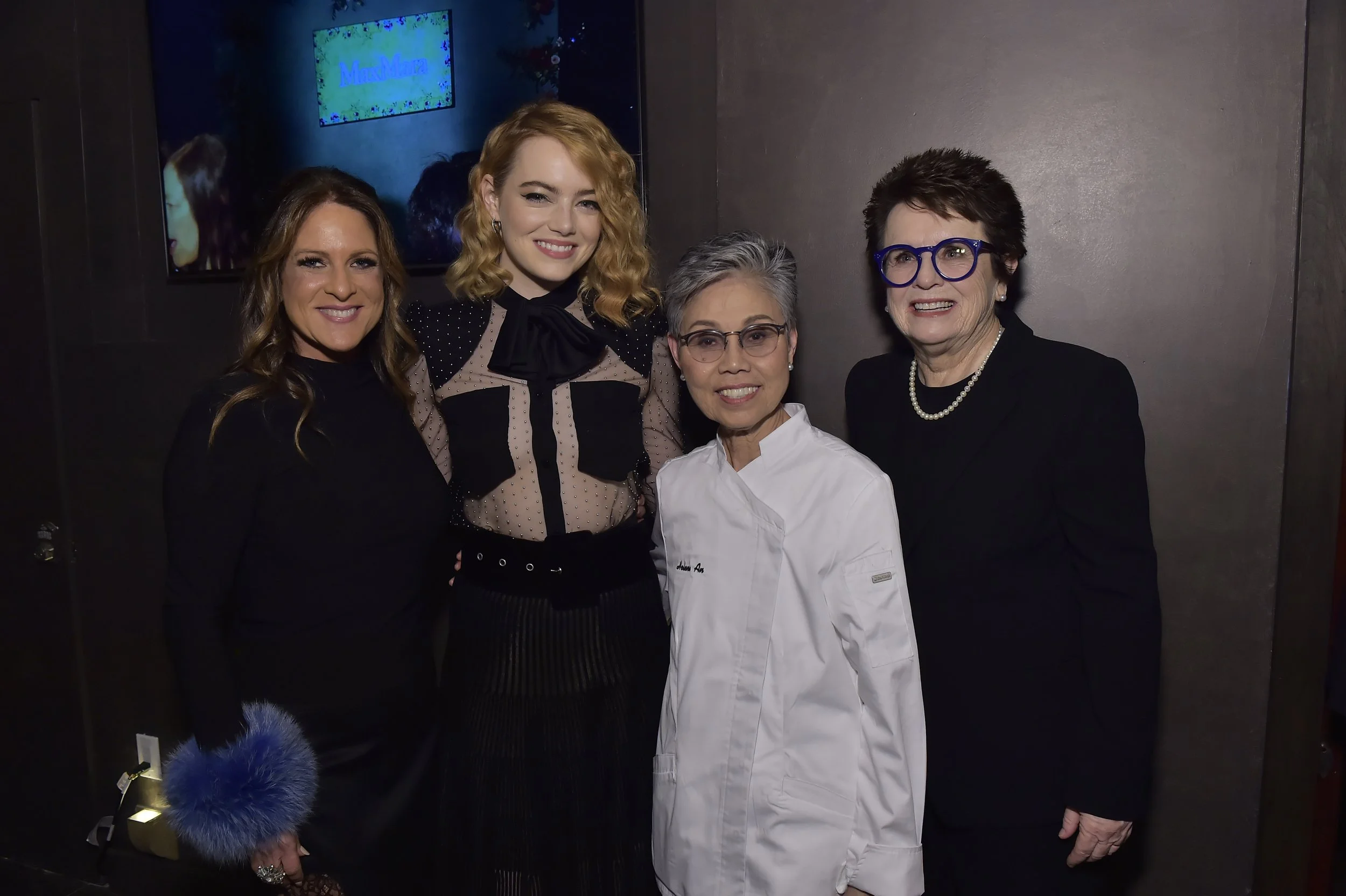 Four women standing together, smiling, posing for a photo at an event. One woman is wearing a white chef's coat, while the others are dressed in black or patterned attire.