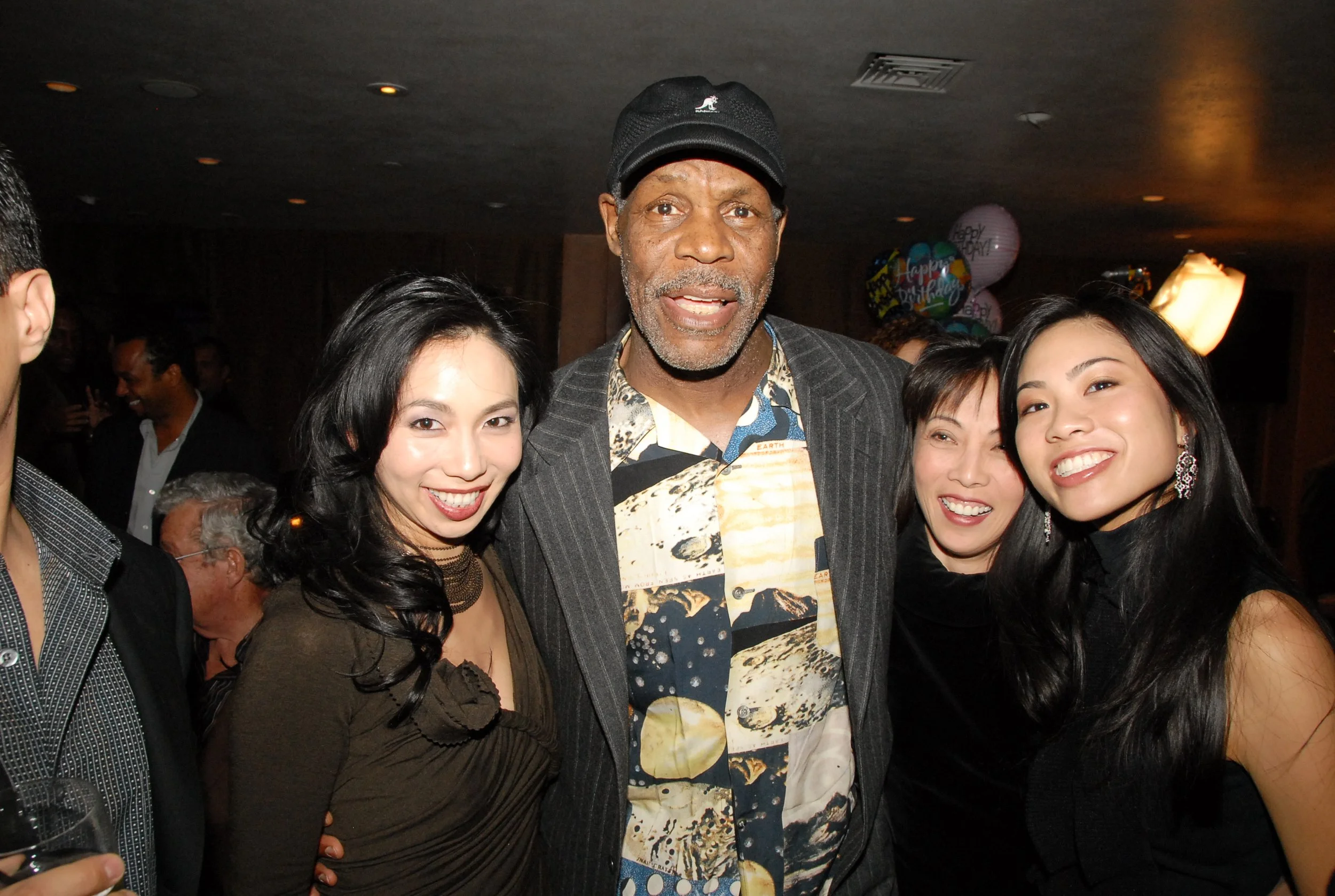 Four people smiling and posing together at a party or celebration, with balloons in the background.