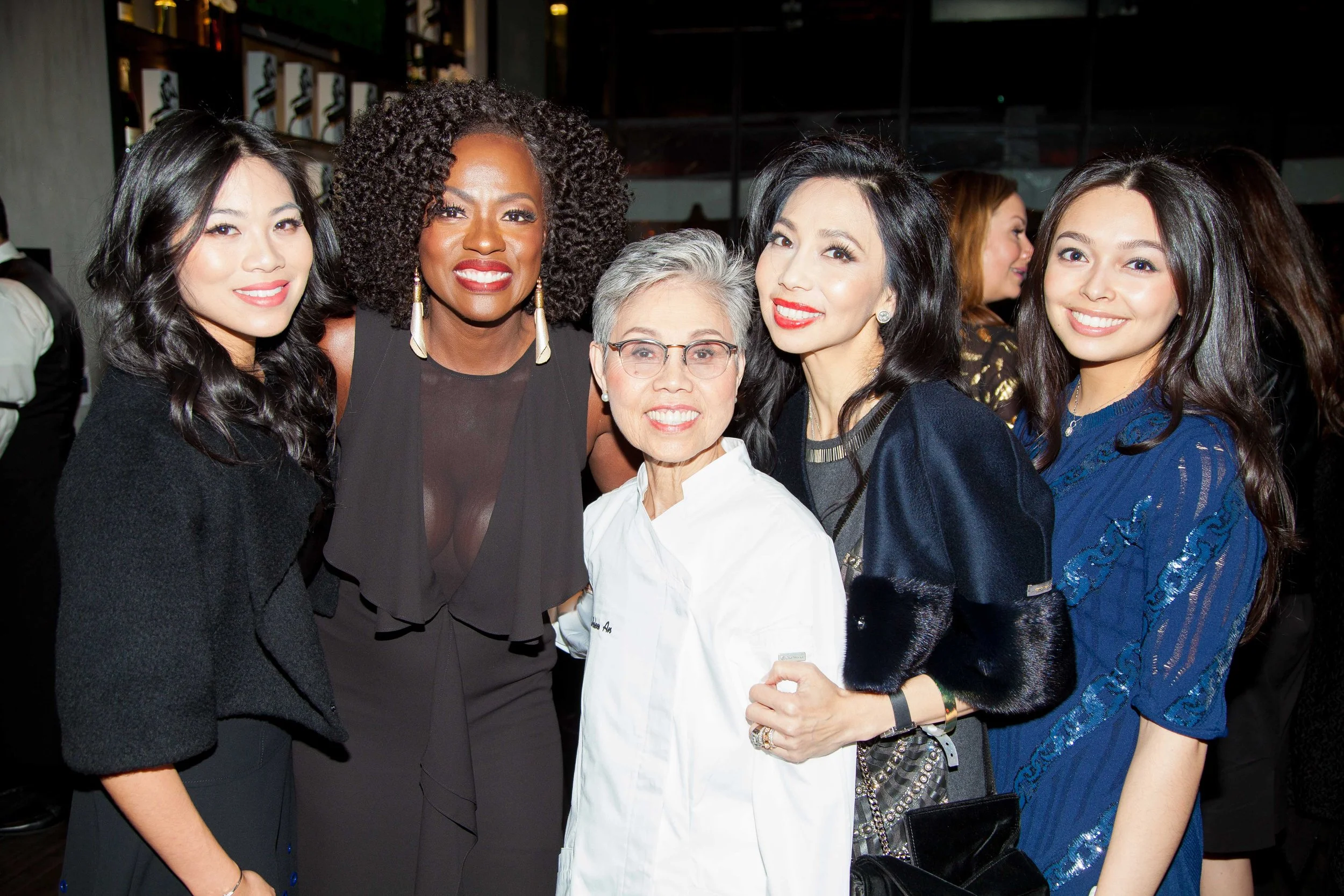 Group of five women at an indoor event, smiling at the camera, dressed in elegant attire.