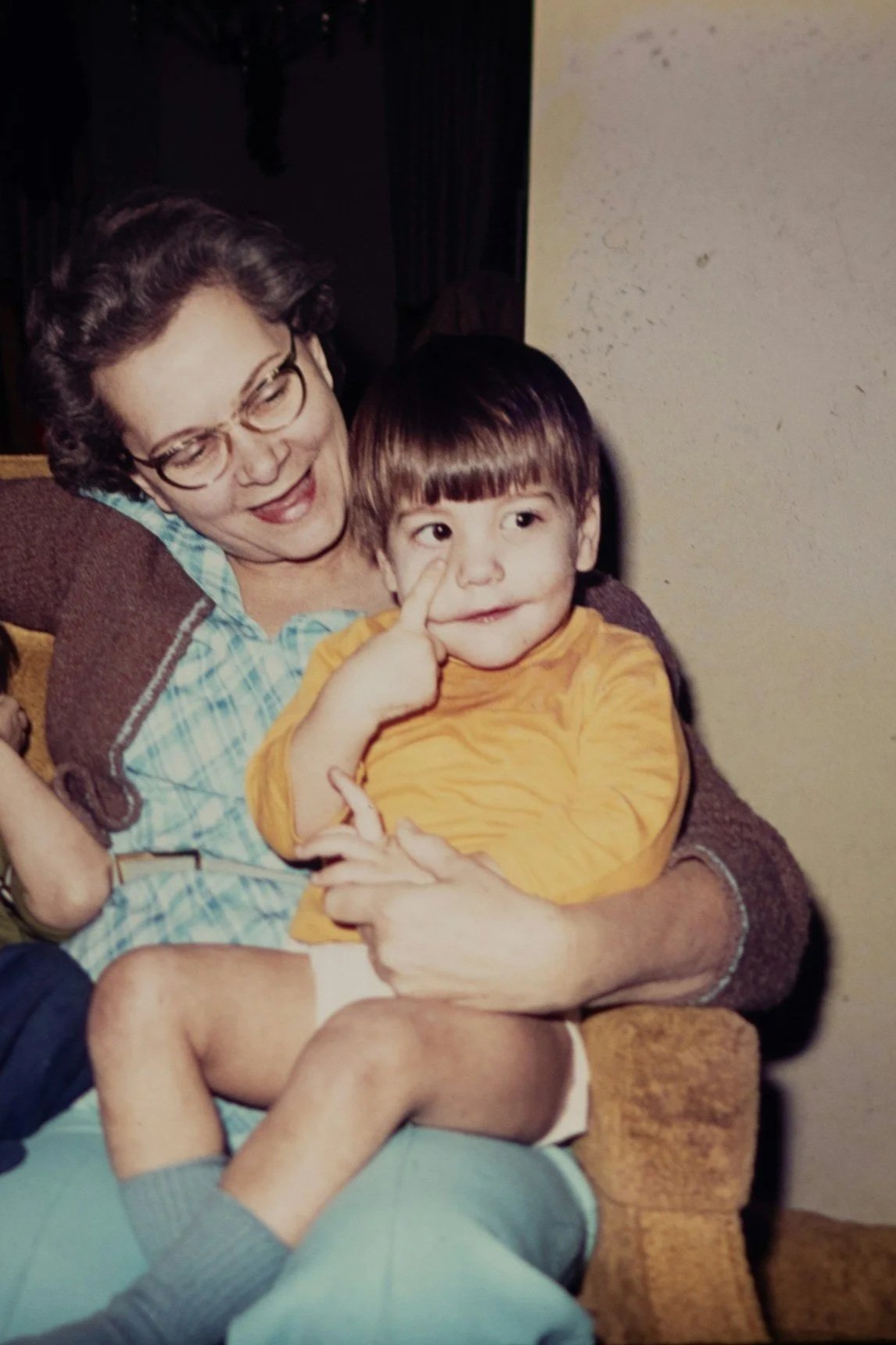 A woman with glasses and curly hair smiling at a young boy with brown hair and wearing a yellow shirt, sitting on her lap.