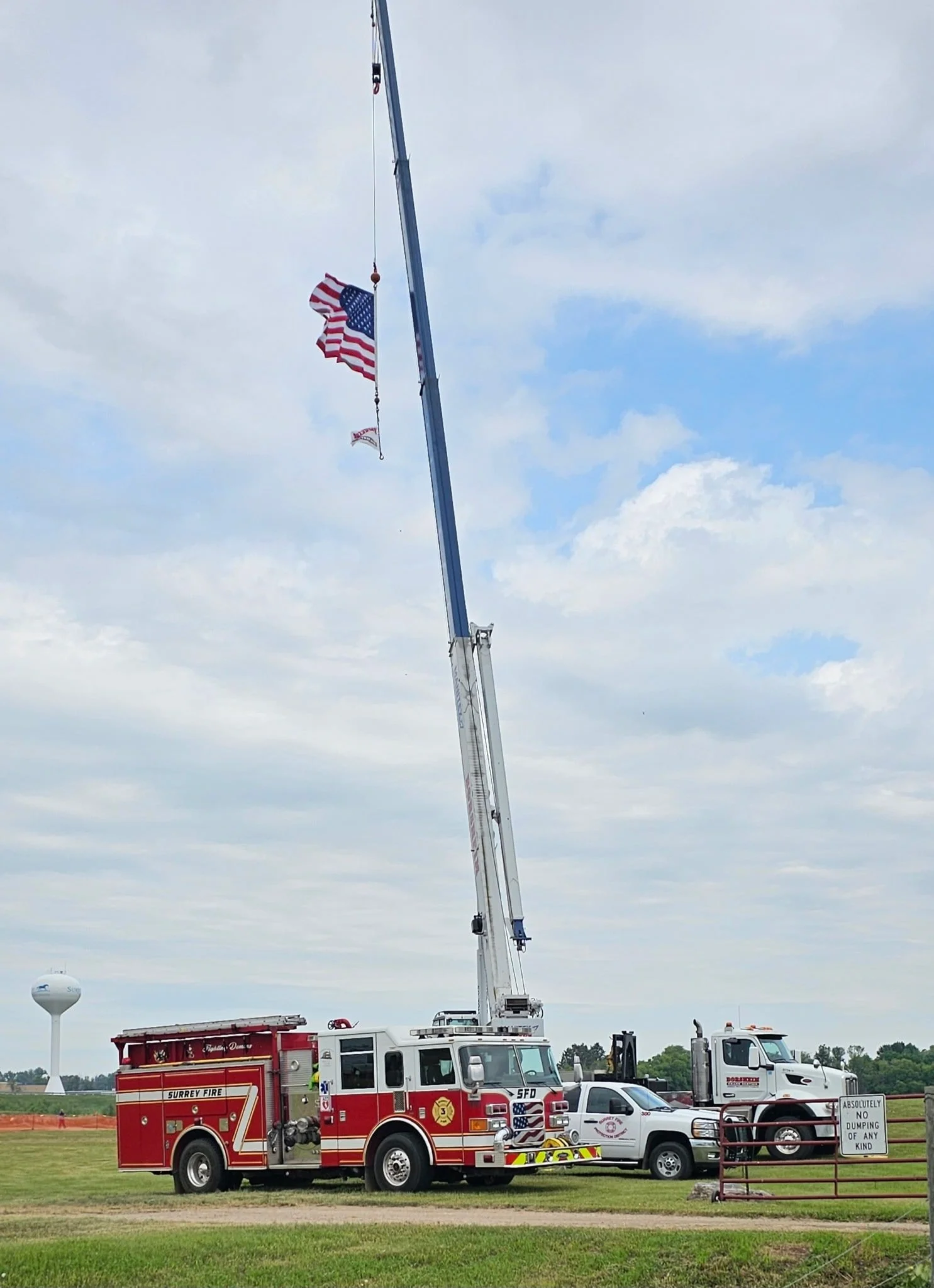 A fire truck with a ladder extended, hoisting an American flag and a smaller flag in a grassy field with a water tower in the background.