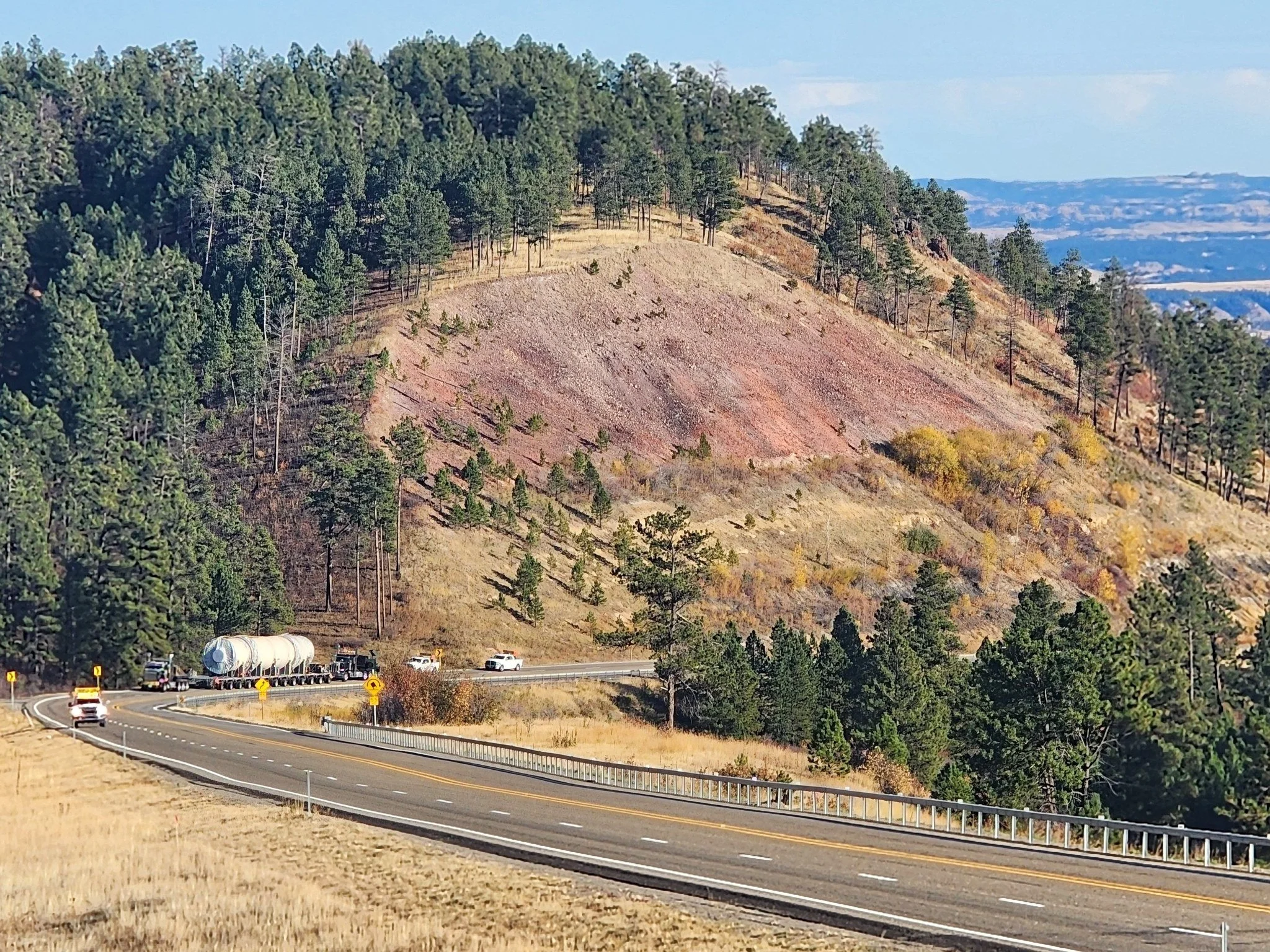 A winding road next to a hillside with scattered trees and gasoline tanks on a flatbed truck, with a backdrop of forested hills and mountains.