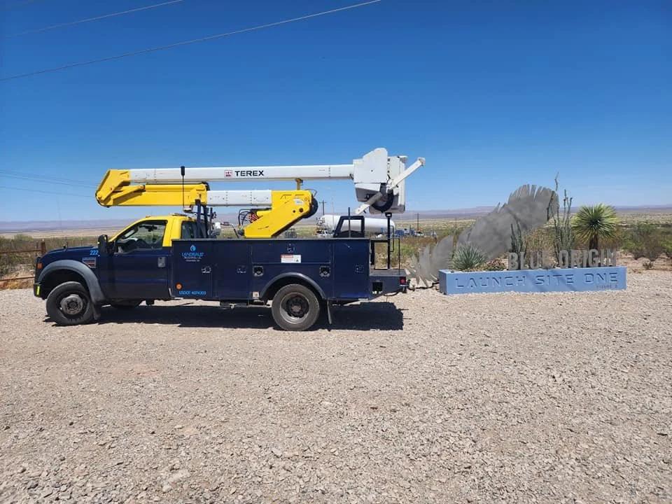A truck with a bucket lift is parked in desert terrain next to a sign that says "Blue Origin Launch Site One," with cactus plants nearby.
