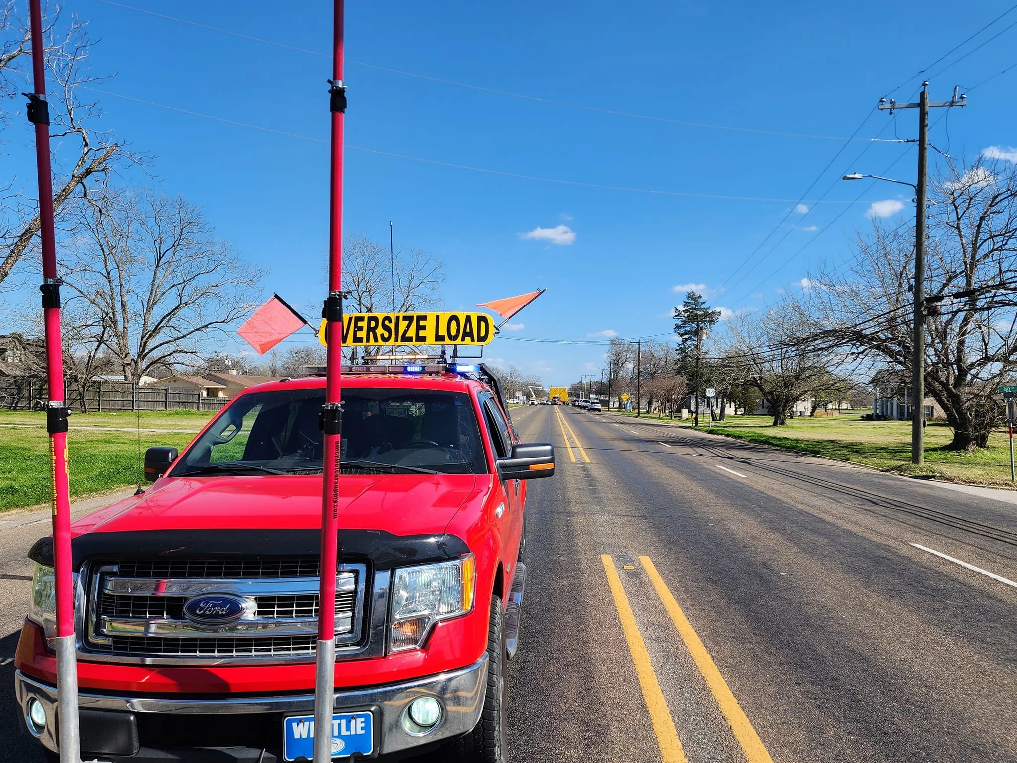 Red pickup truck with a yellow sign reading 'OVERSIZE LOAD' on top, blocking the road with orange and pink flags attached, on a clear day with a blue sky and trees along the street.