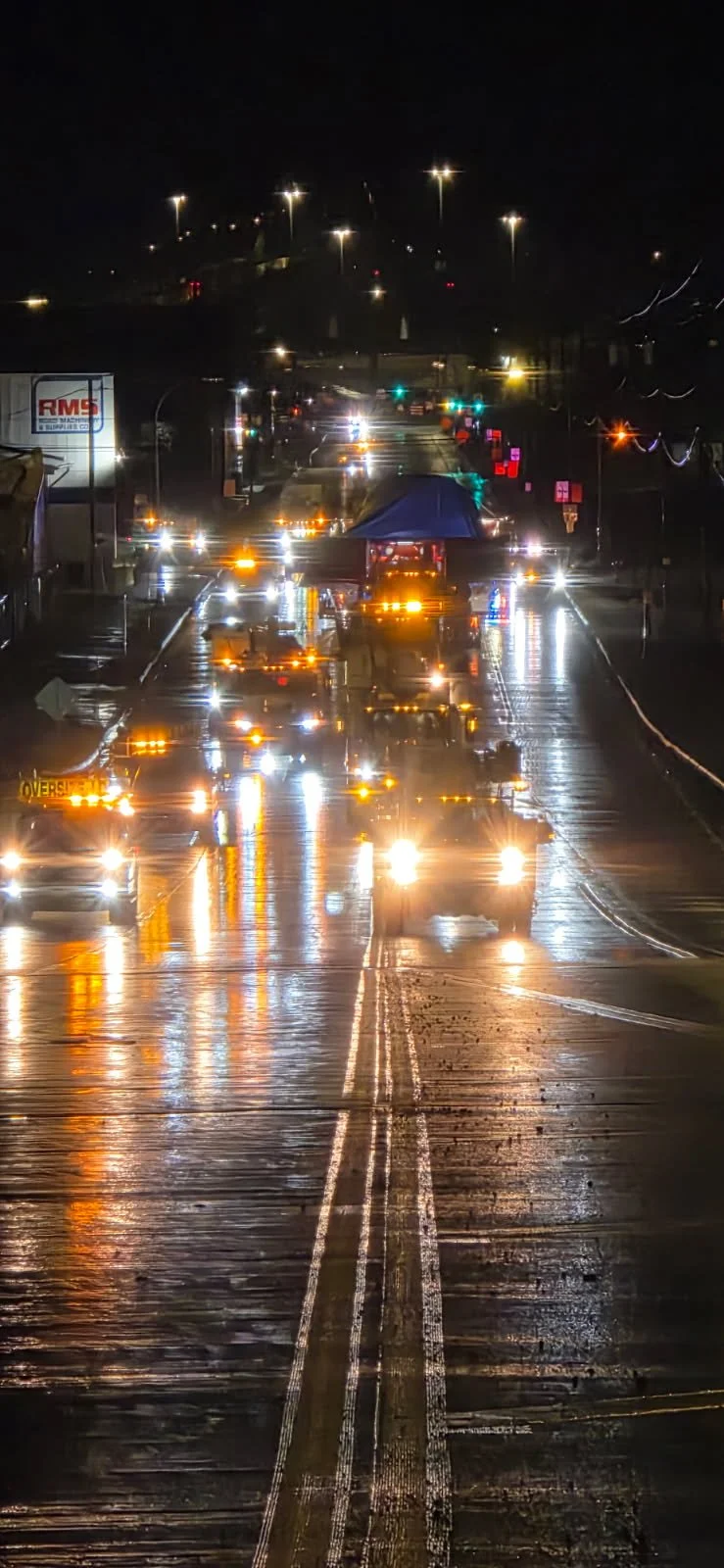 Nighttime street scene with wet road reflecting streetlights and vehicle headlights, cars, and a temporary windbreak or tent set up on the street.