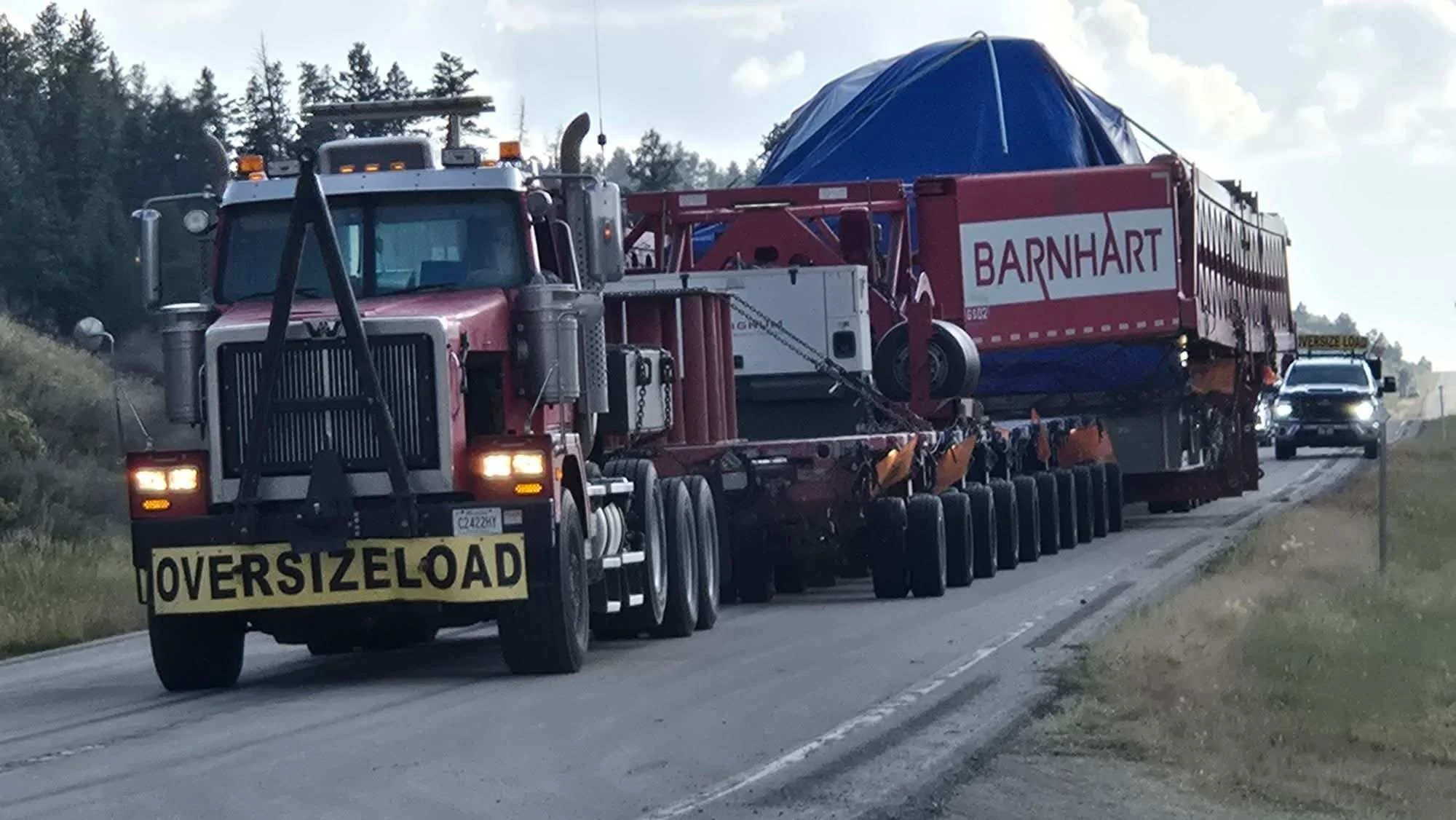 Large oversize load carrying a barnart structure on a flatbed truck, followed by a police car, on a two-lane rural road with trees and cloudy sky in the background.