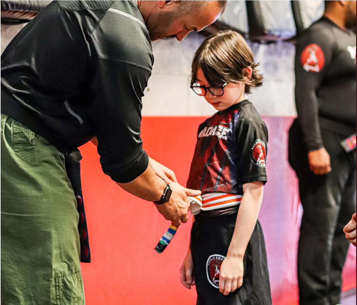 A martial arts instructor helps a young boy put on a martial arts belt during a belt ceremony, with another instructor standing in the background.