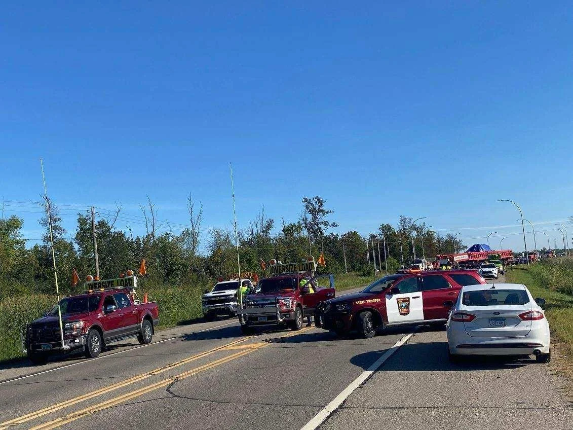 Multiple emergency vehicles, including state trooper and oversized load escort trucks, are stopped on a rural road with trees in the background under a clear blue sky.
