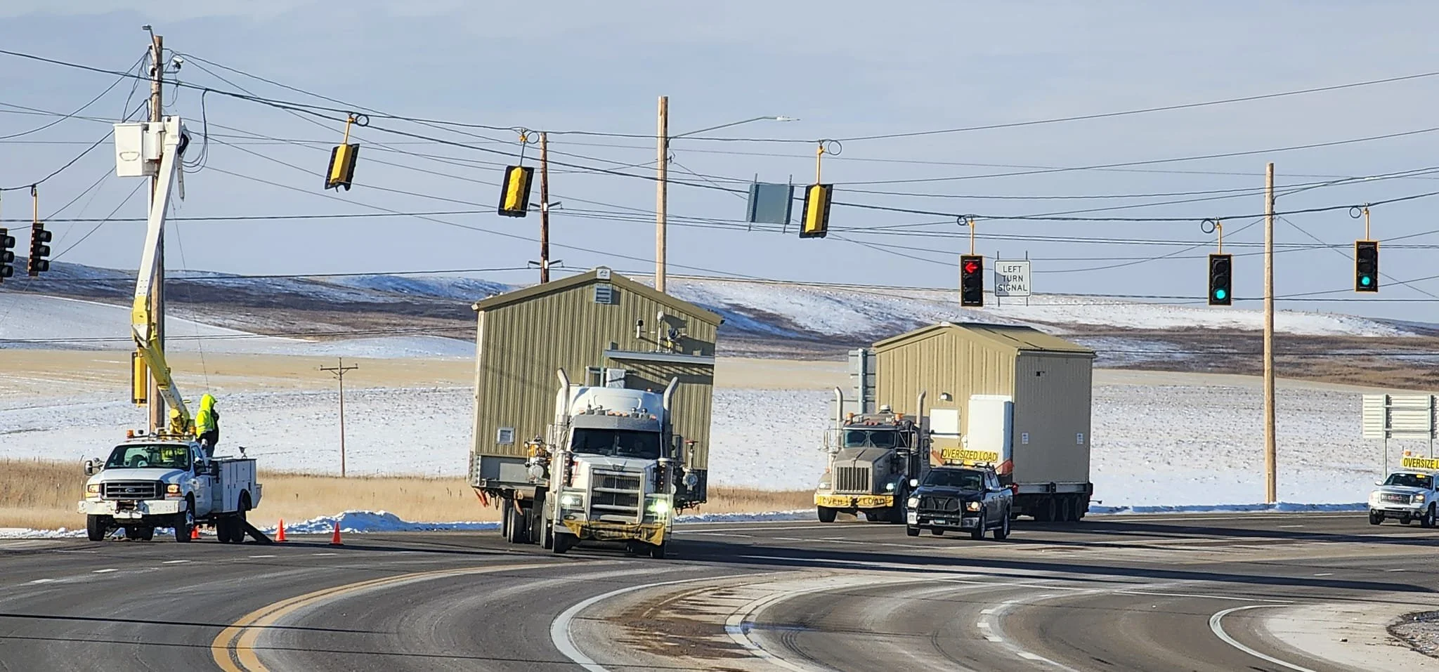 Road construction scene with utility trucks and workers, some workers are in a bucket lift, snow on the ground, traffic lights and signs, and a rural landscape in the background.