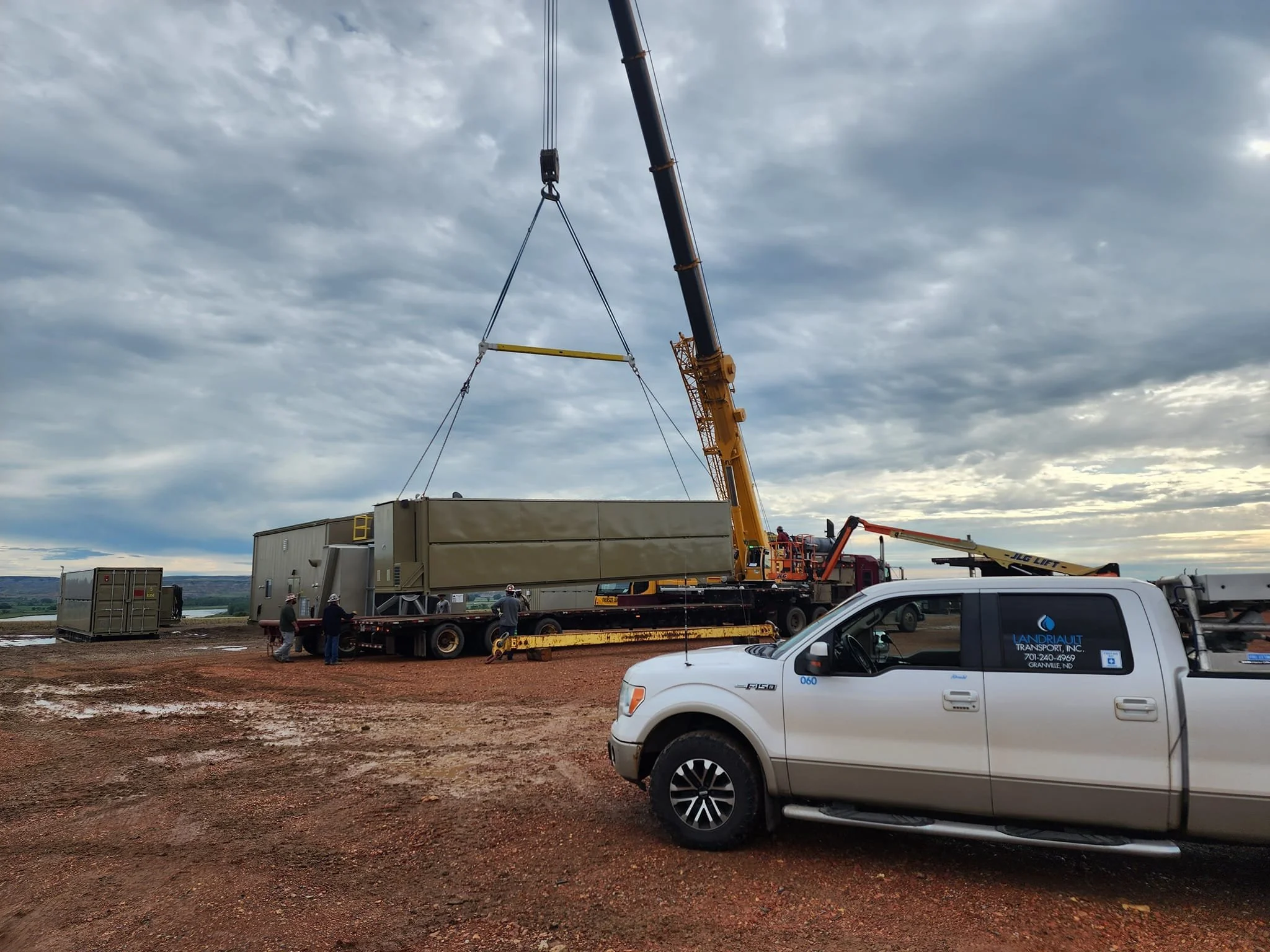 A crane lifting a large beige industrial container onto a flatbed truck at a construction site on a cloudy day with a few people guiding the load.