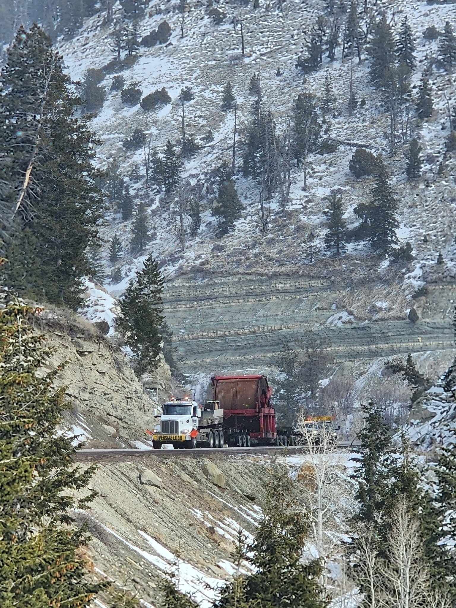 A large truck with an oversize load banner is driving through a snowy mountain landscape on a narrow road surrounded by trees and rocky slopes.