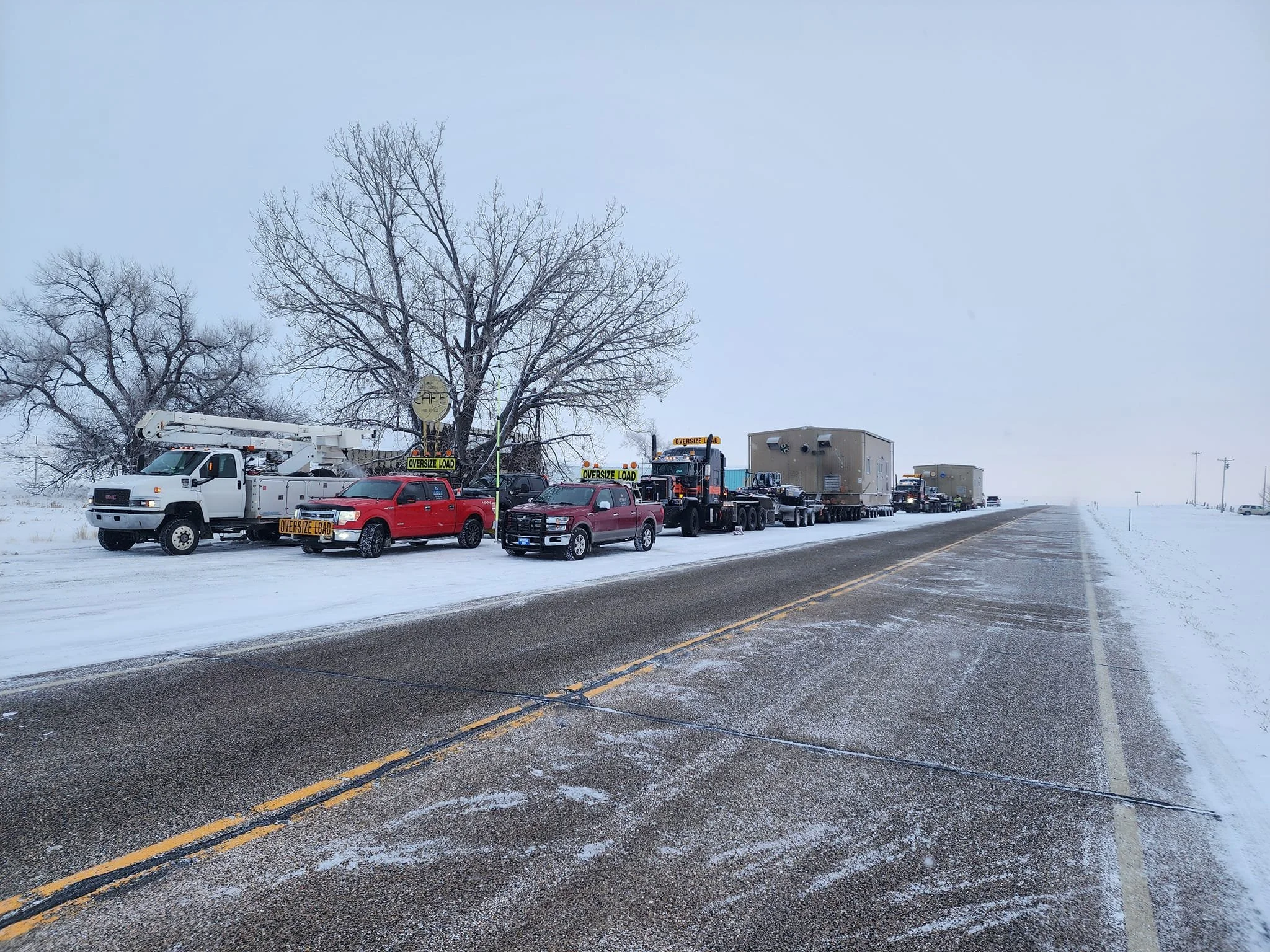 Line of vehicles, including trucks and utility vehicles, parked on the side of a snowy road with trees in the background.