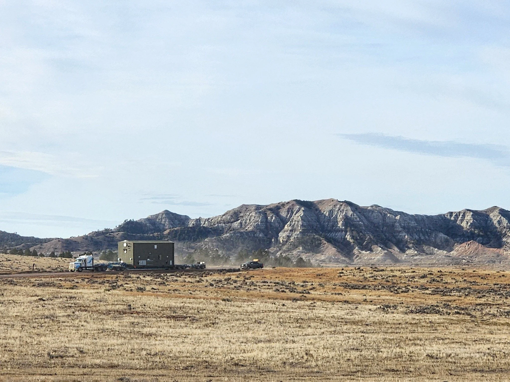A flat, open desert landscape with dry grass, distant mountains, and a few vehicles, including a white truck, a darker vehicle, and a large dark structure on a trailer.