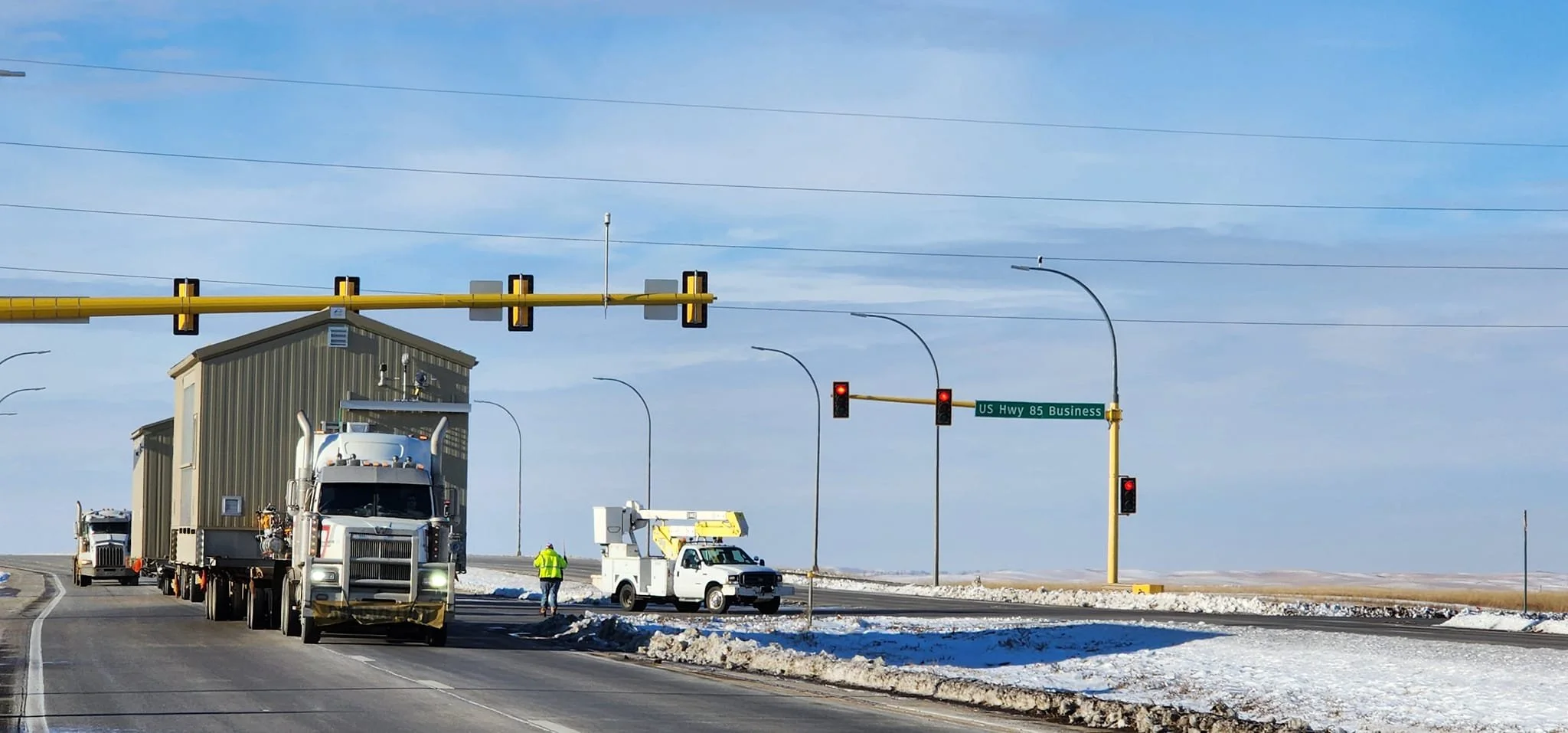 Semi-truck hauling a small building on a snowy highway, with a worker and utility truck nearby, and traffic lights at an intersection under a blue sky.
