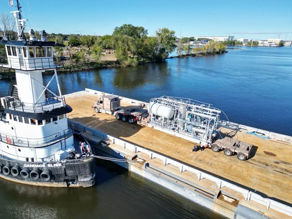 A large ship docked along a river with trees and industrial buildings in the background. The ship's deck has industrial equipment and a trailer.