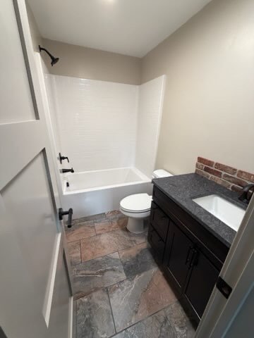 A small bathroom with a bathtub and shower, a toilet, and a dark wood vanity with a sink, brick backsplash, and gray countertop.