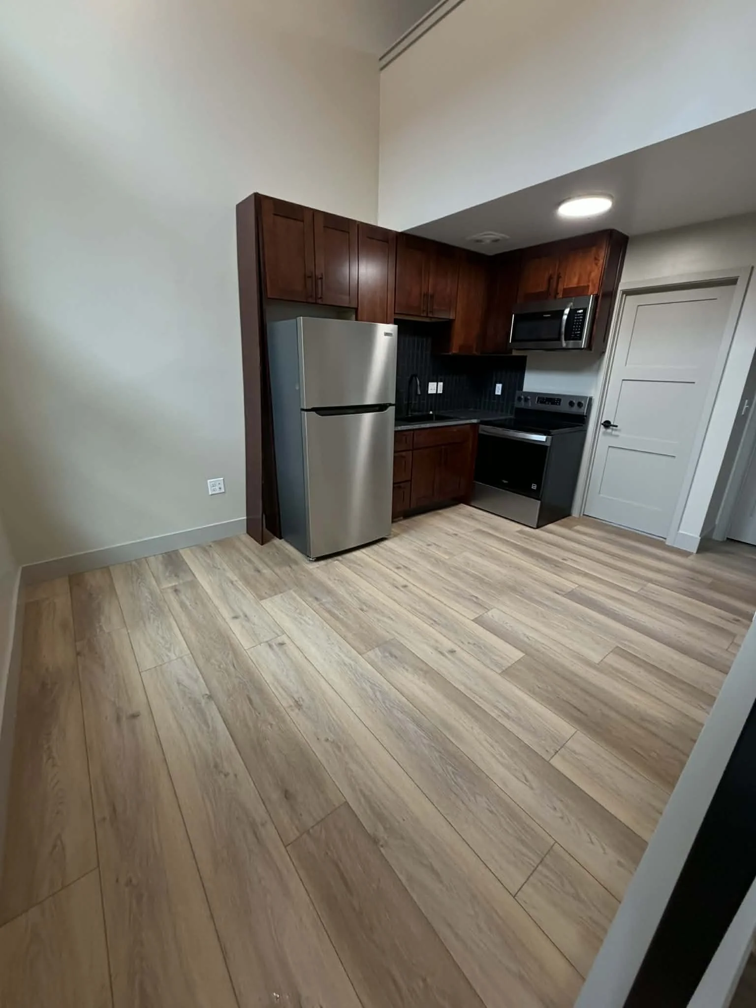 Kitchen with wooden cabinets, stainless steel refrigerator, microwave, and oven, with light wood flooring and a white wall on the left.