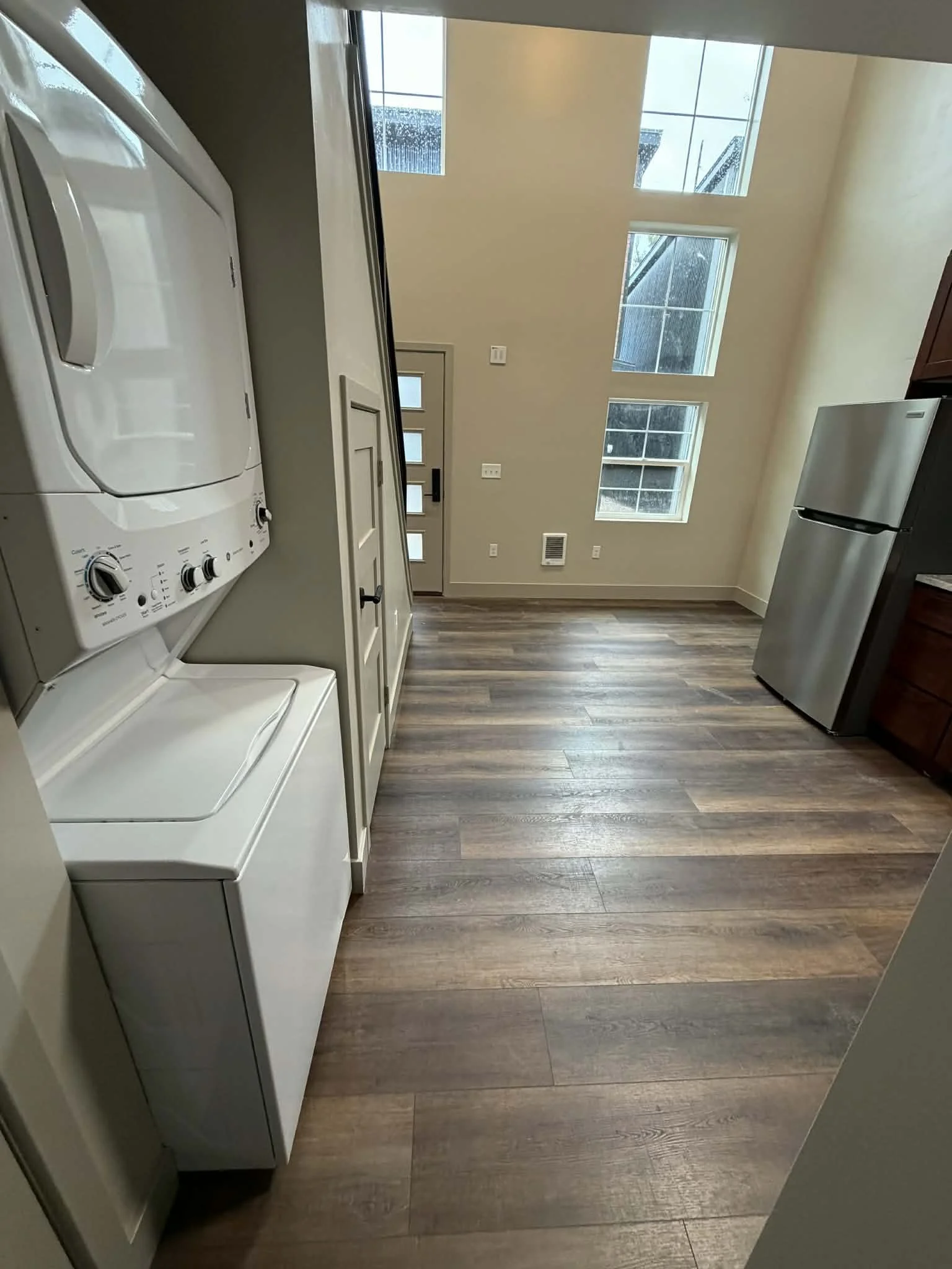 Laundry room with stacked washer and dryer on the left side, hardwood floor, and a large window wall letting in natural light.