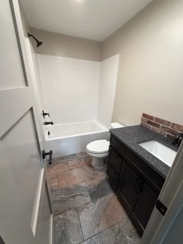 Small bathroom with bathtub, toilet, and dark vanity with a sink, brick accent wall, and gray tiled floor.