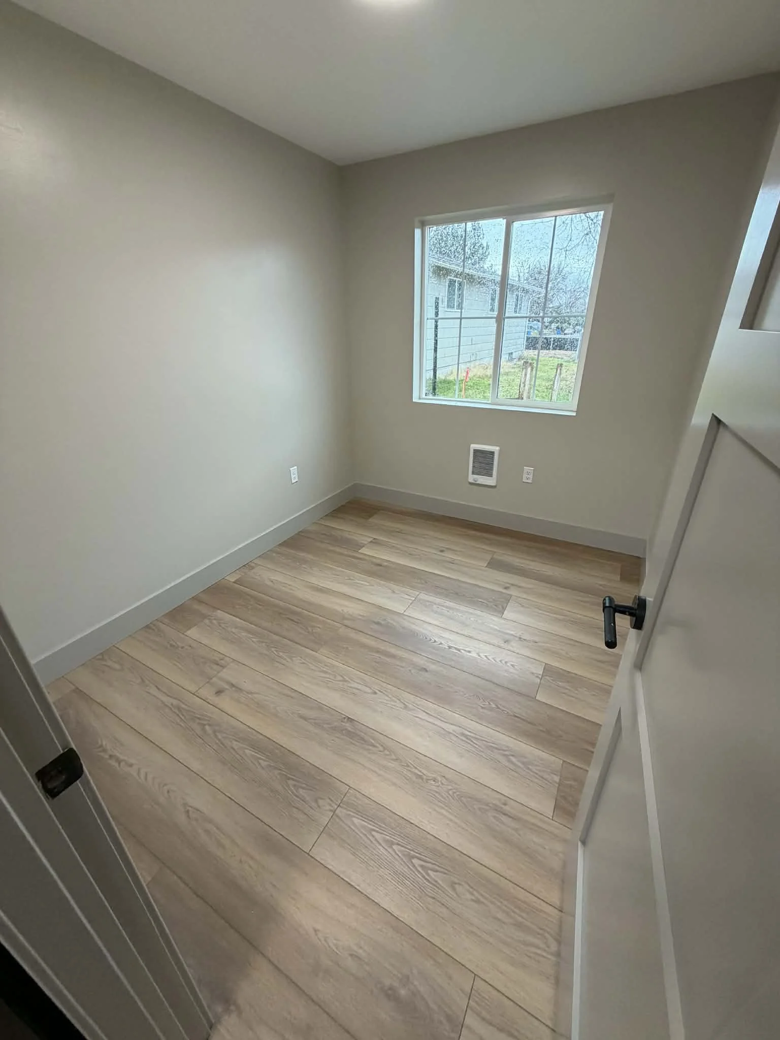 Empty room with beige walls, a window showing an outdoor yard, light wood flooring, and a white door partially open.