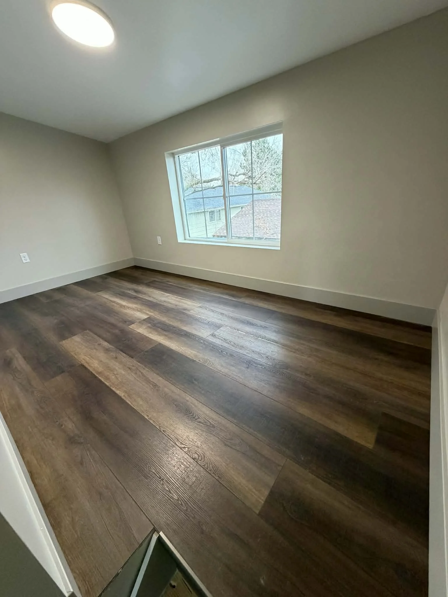Empty room with wood flooring, white walls, a window showing neighborhood trees and rooftops, and a ceiling light.