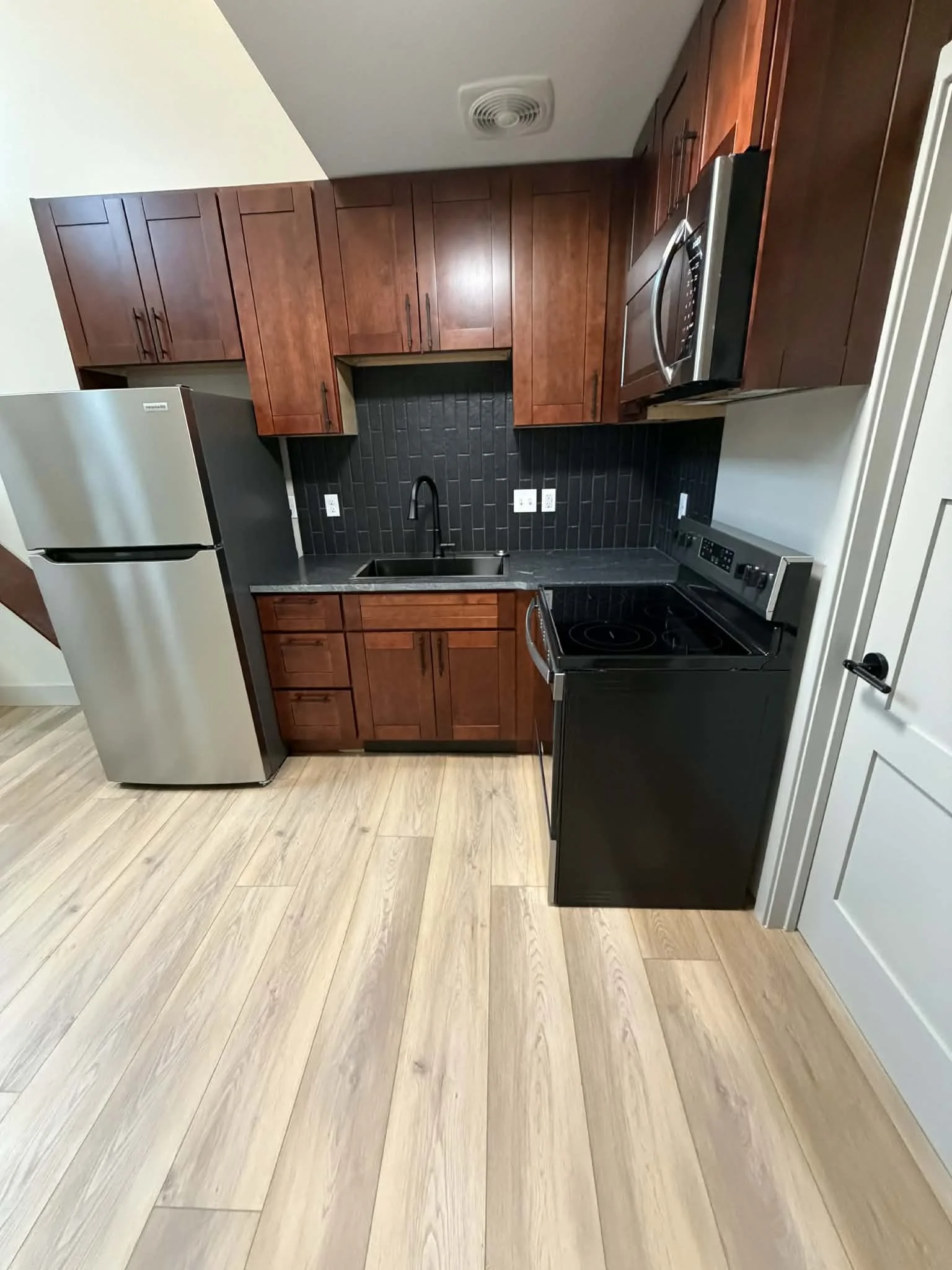 A modern kitchen with dark wood cabinets, a black backsplash, stainless steel refrigerator and microwave, and black stove and sink, all on light wood flooring.