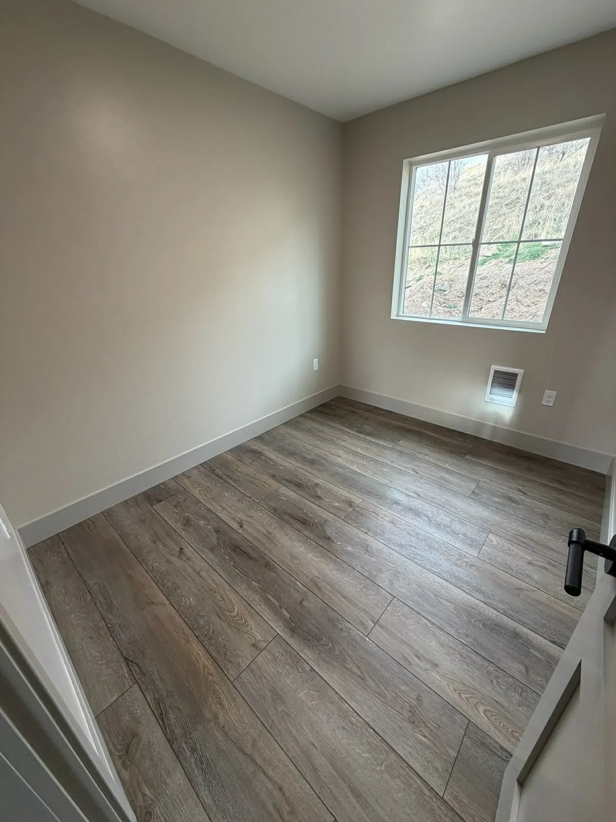 Empty room with beige walls, wood-like flooring, a square window with a scenic outdoor view, and a small vent near the floor.
