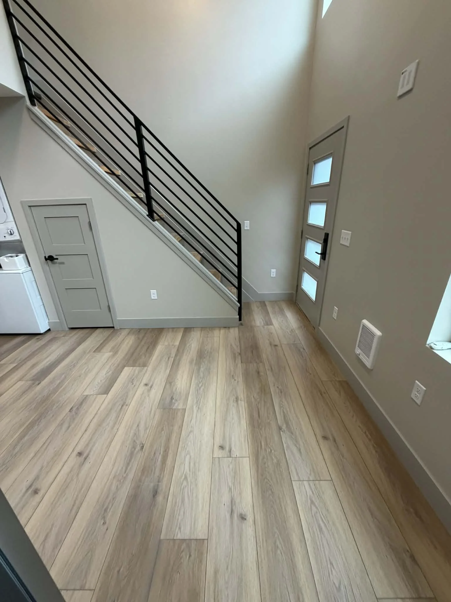 Interior of a modern house with light hardwood flooring, a staircase with black metal railing leading to the upper floor, a gray door under the stairs, a front door with windows, small wall-mounted heater, and multiple electrical outlets on light-colored walls.