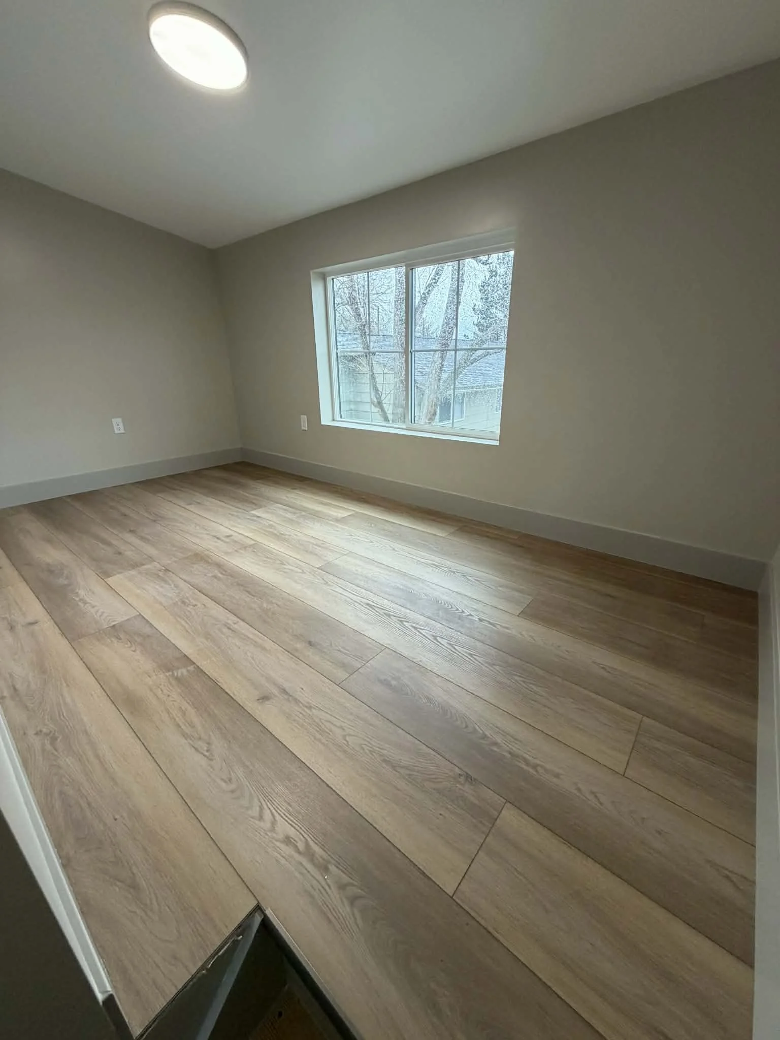 Empty room with hardwood floors, beige walls, a large window, and a ceiling light.