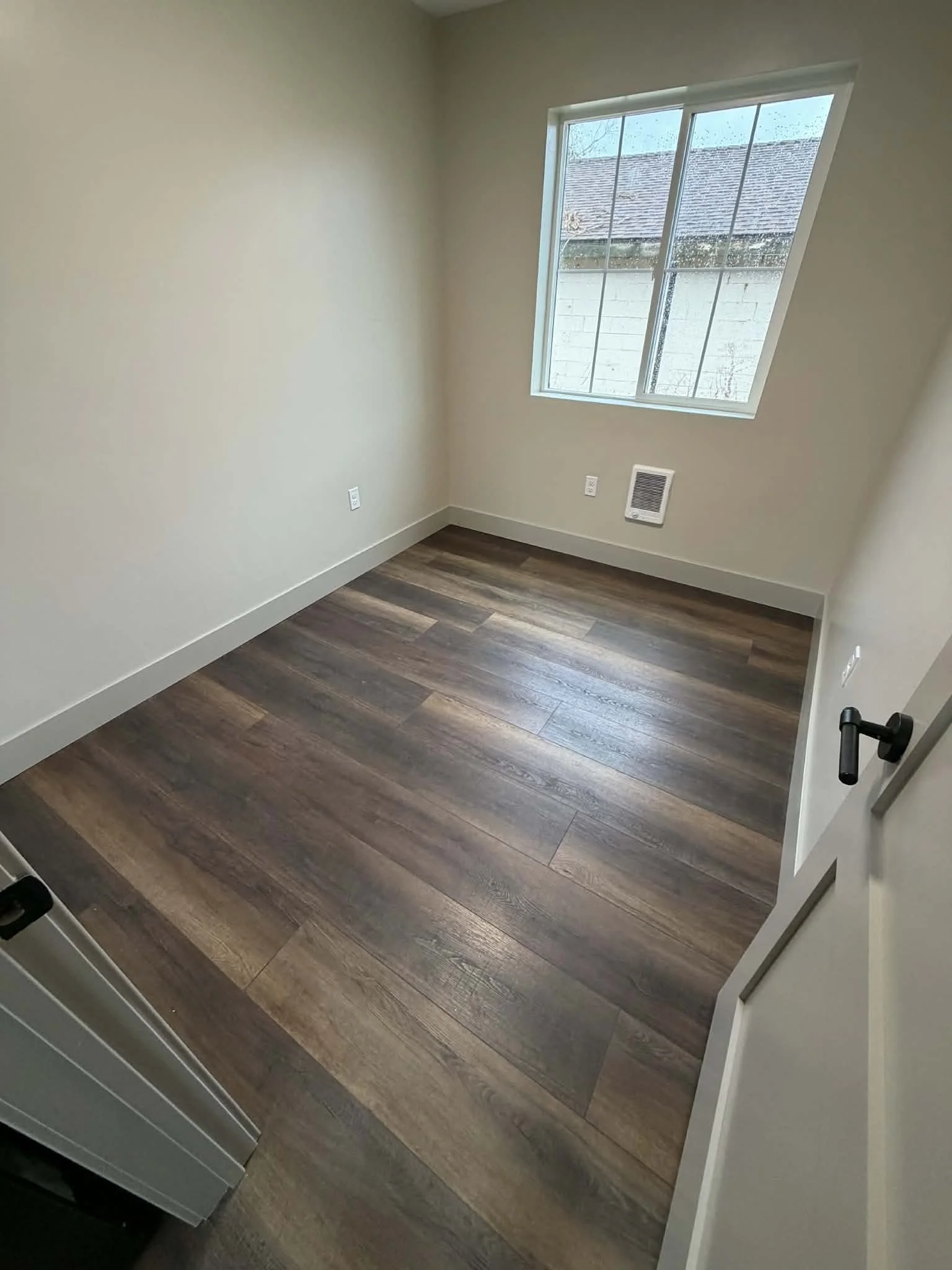 Empty room with beige walls, wooden flooring, a window with rain outside, a vent on the wall, and a partially visible door.