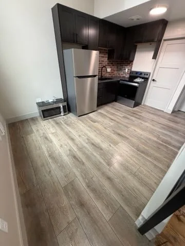 Kitchen with dark cabinets, stainless steel refrigerator, black stove, a brick backsplash, and wood flooring.
