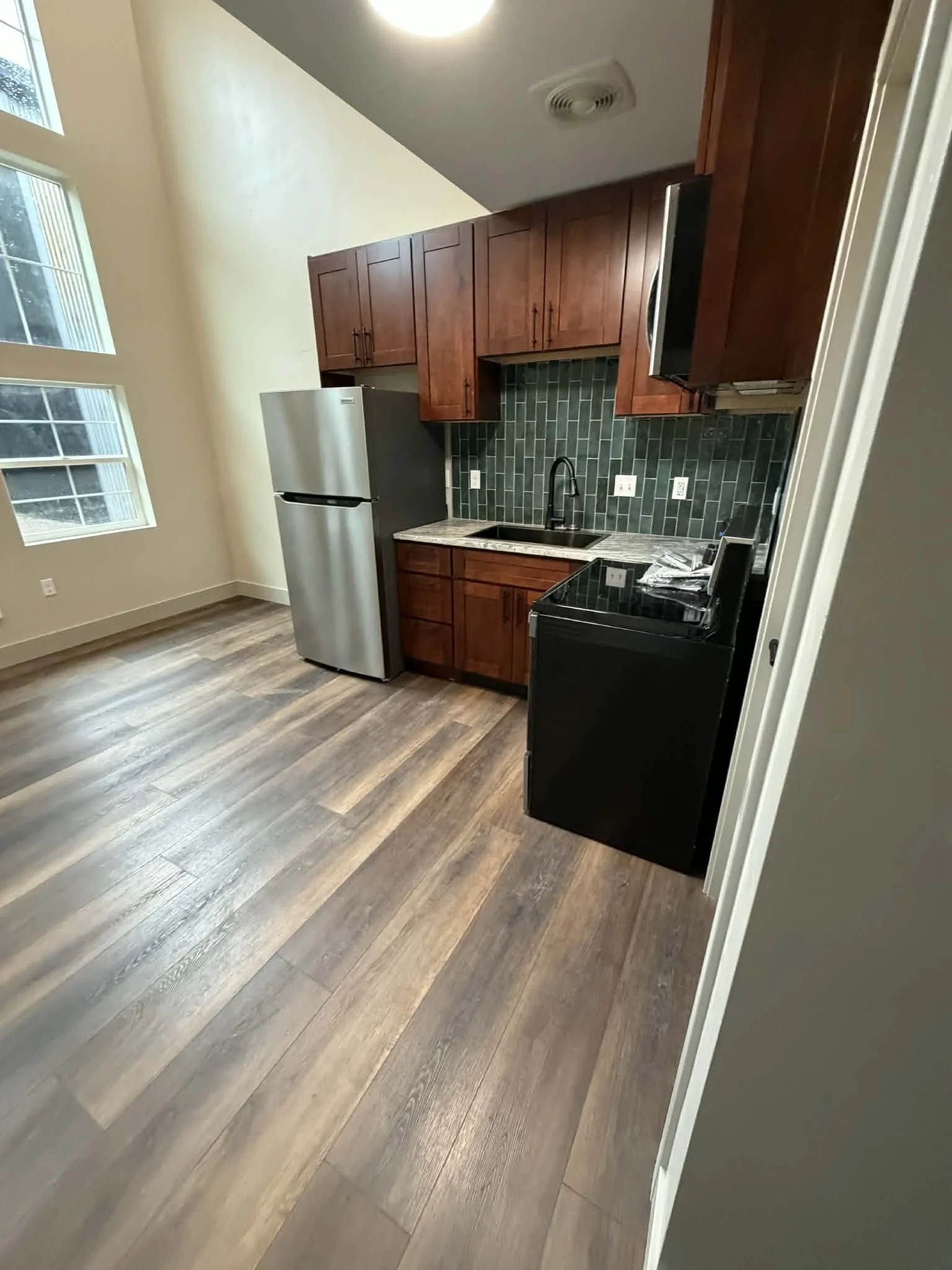 Interior view of a modern kitchen with wooden cabinets, black stove, and stainless steel refrigerator next to a green tiled backsplash and a black sink under a high ceiling with large windows on the left side.