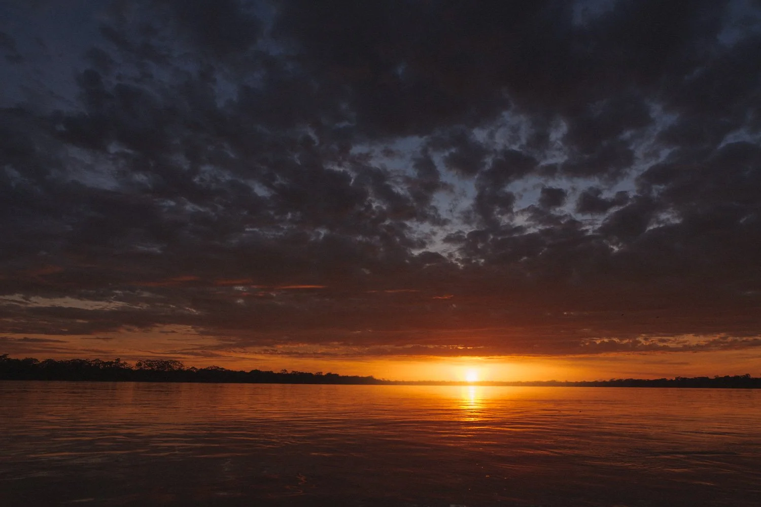 Sunset over a calm river with dark clouds in the sky and a faint orange glow on the horizon.