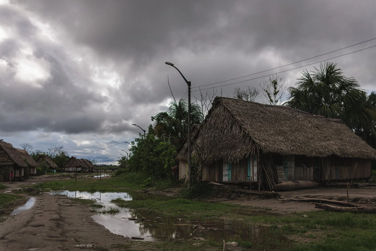 A muddy street lined with thatched homes under a stormy sky in the Peruvian Amazon.
