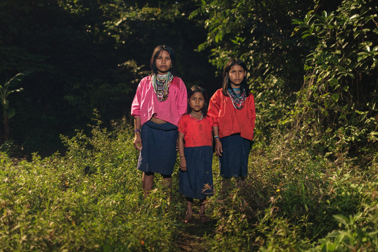 Three young sisters in traditional clothing stand together in the dense greenery of the Amazon rainforest.