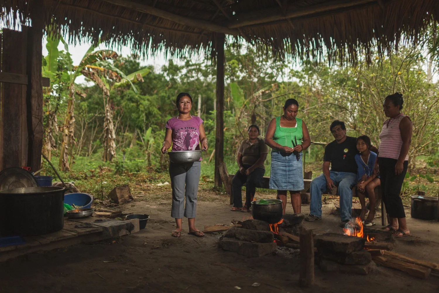 A family prepares a meal over an open fire inside a traditional Amazonian kitchen shelter.
