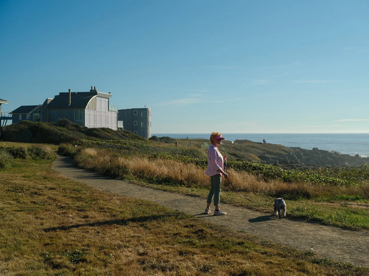 A woman walks her dog along a coastal path near Overleaf Lodge in Yachats, Oregon, with ocean views and seaside hotel rooms in the background.