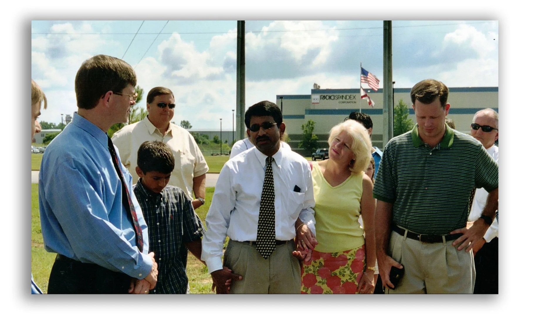 Group of people standing outdoors during daytime, engaged in conversation, with a building, American flags, and a blue sky with clouds in the background.
