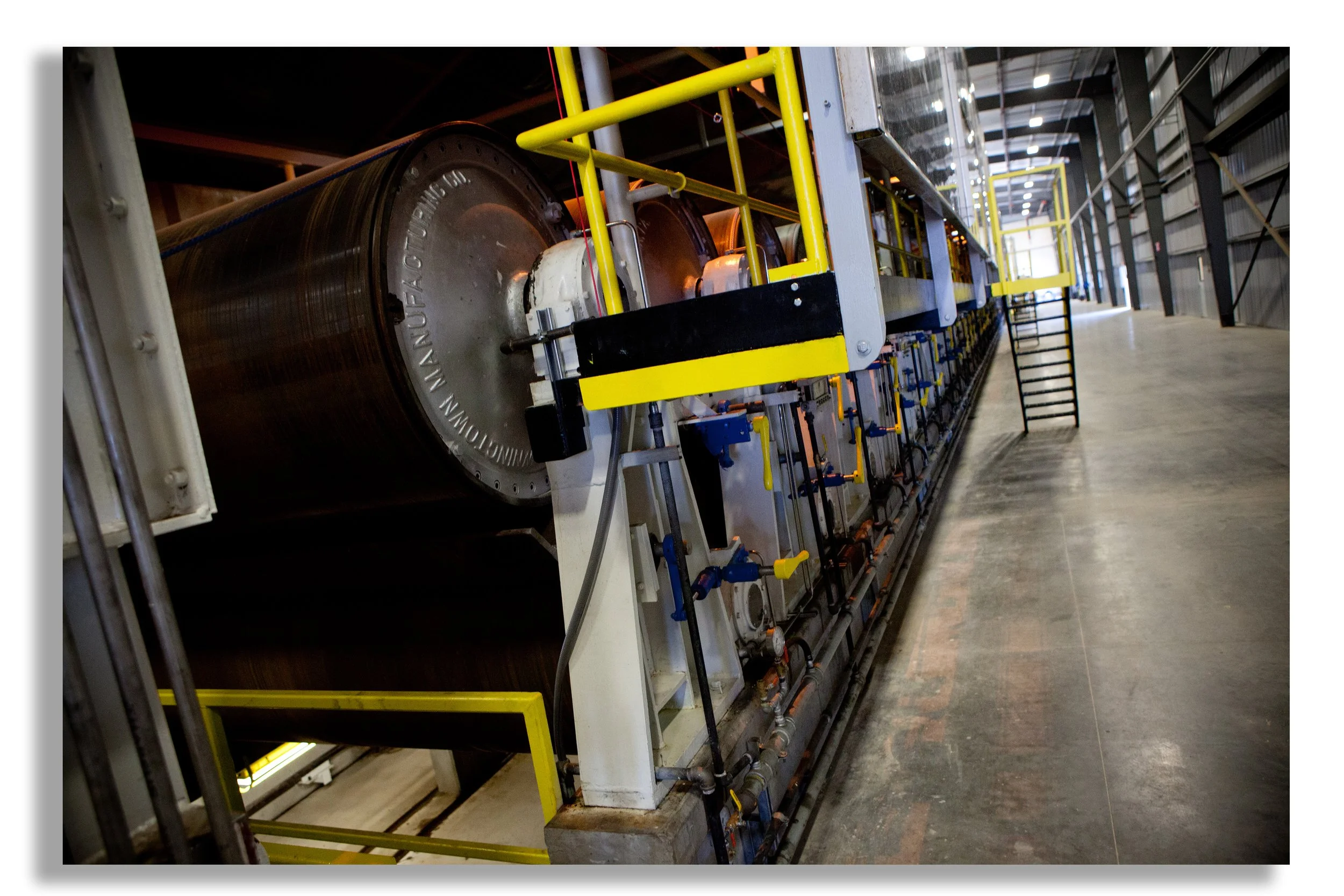 Long industrial machine with large metallic cylinders, yellow safety railings, and control panels inside a warehouse.