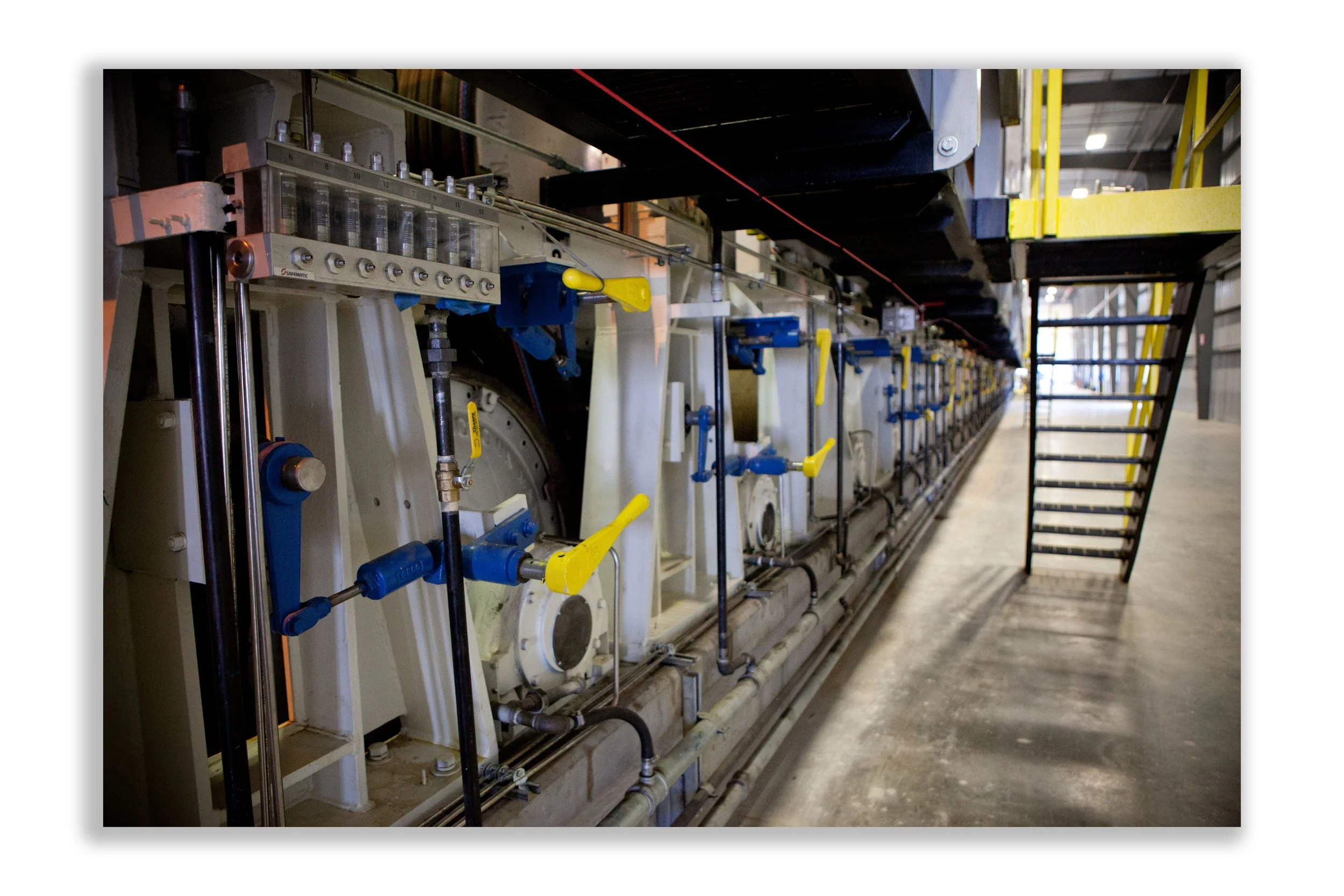 Industrial machinery with pipes, valves, and levers in a factory setting, with stairs and a walkway on the right.