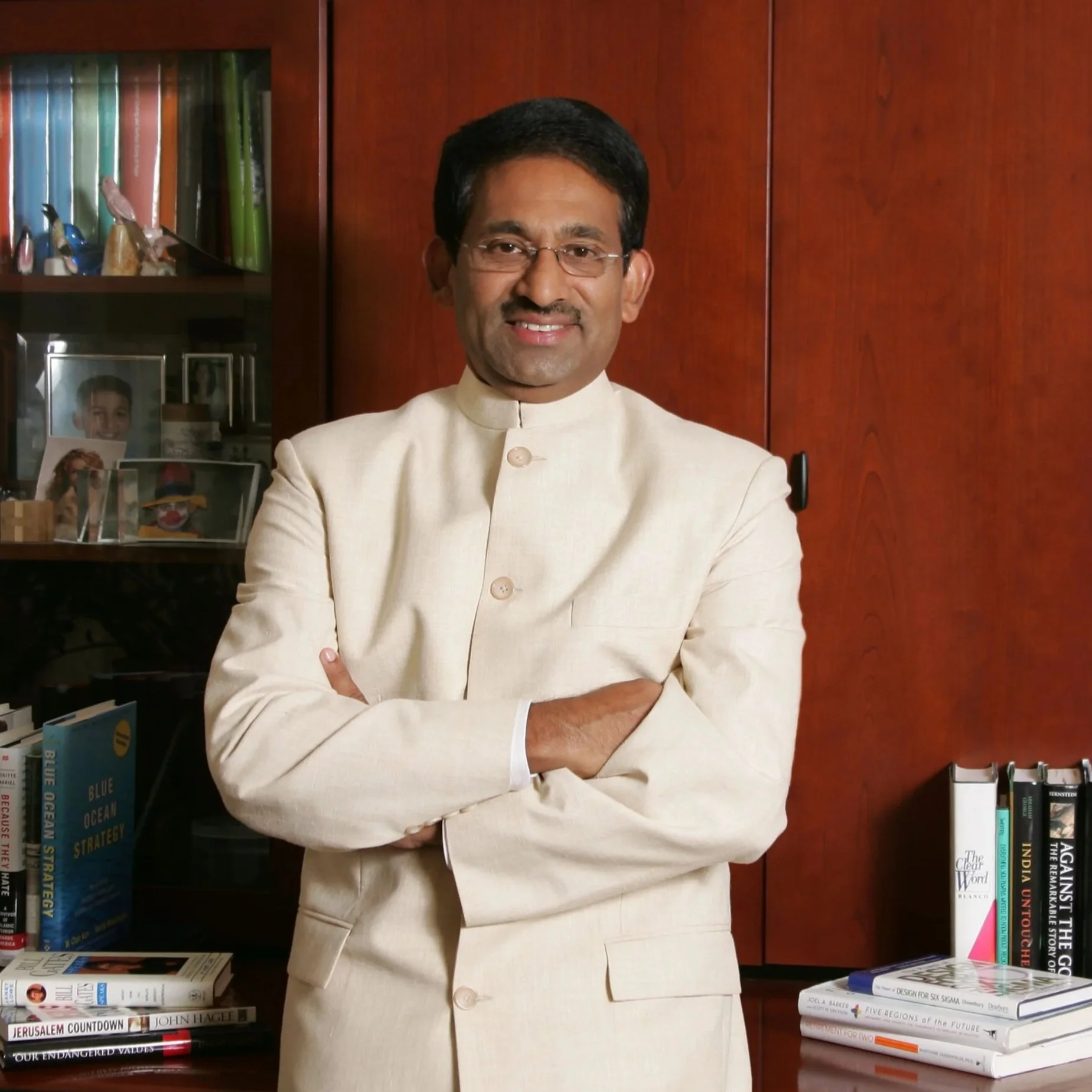 A man wearing traditional Indian attire standing with crossed arms in front of a bookshelf and wooden cabinet in a study or office.
