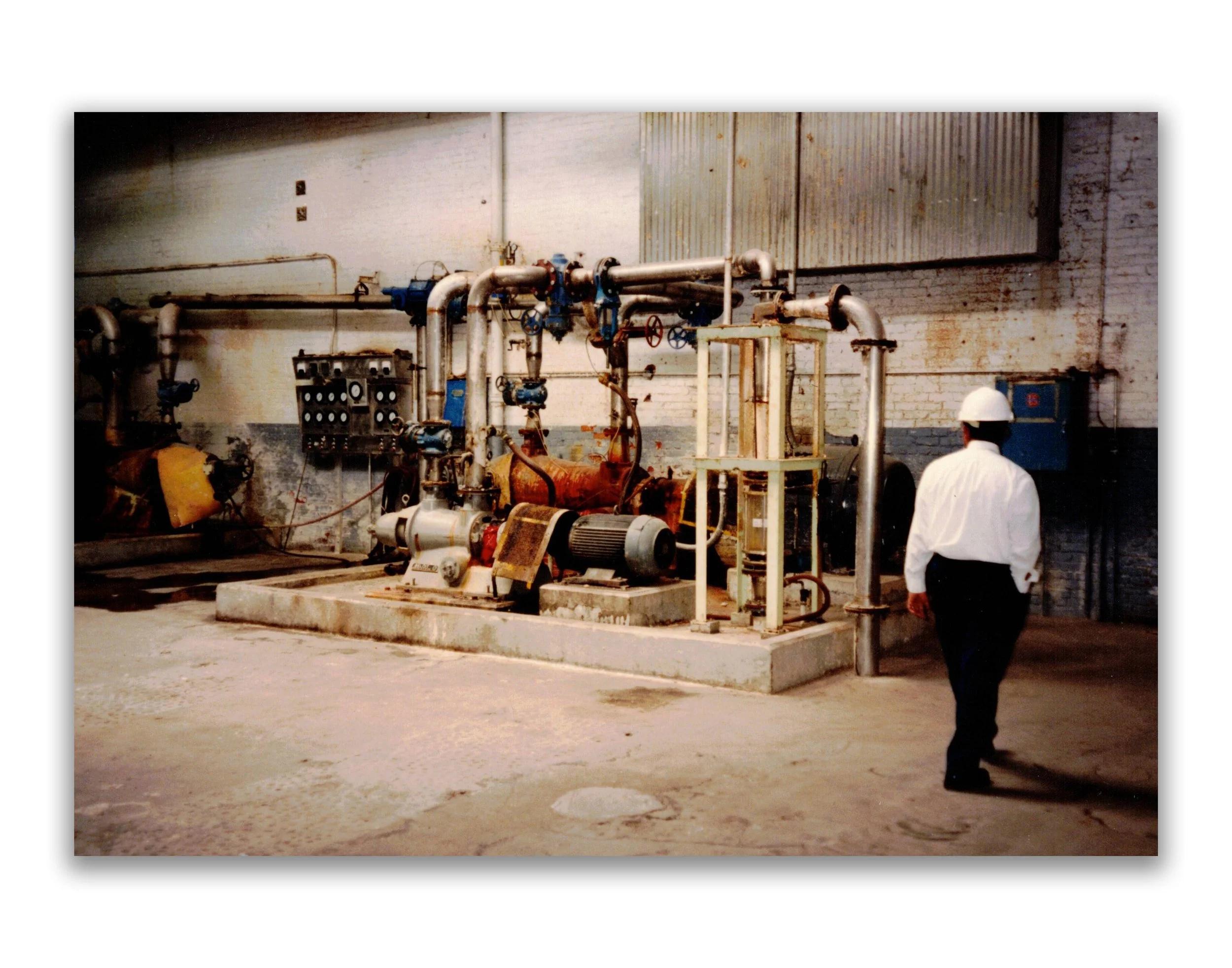 A man wearing a white safety helmet and white shirt stands in an industrial facility with pipes, valves, and equipment on a concrete platform.