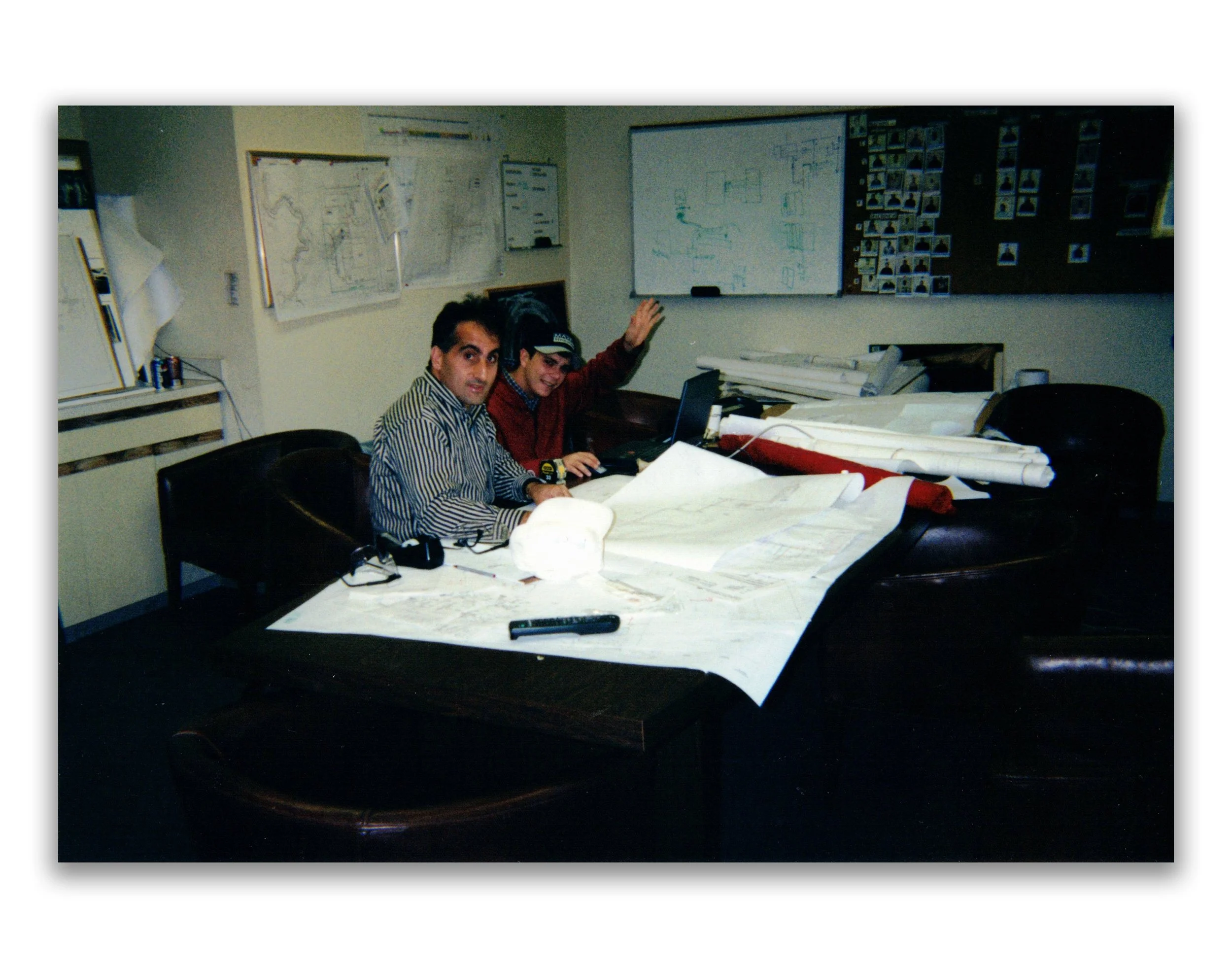 Two men sitting at a cluttered conference table with rolled-up blueprints and a large sheet of paper, in an office with maps and a whiteboard on the wall.