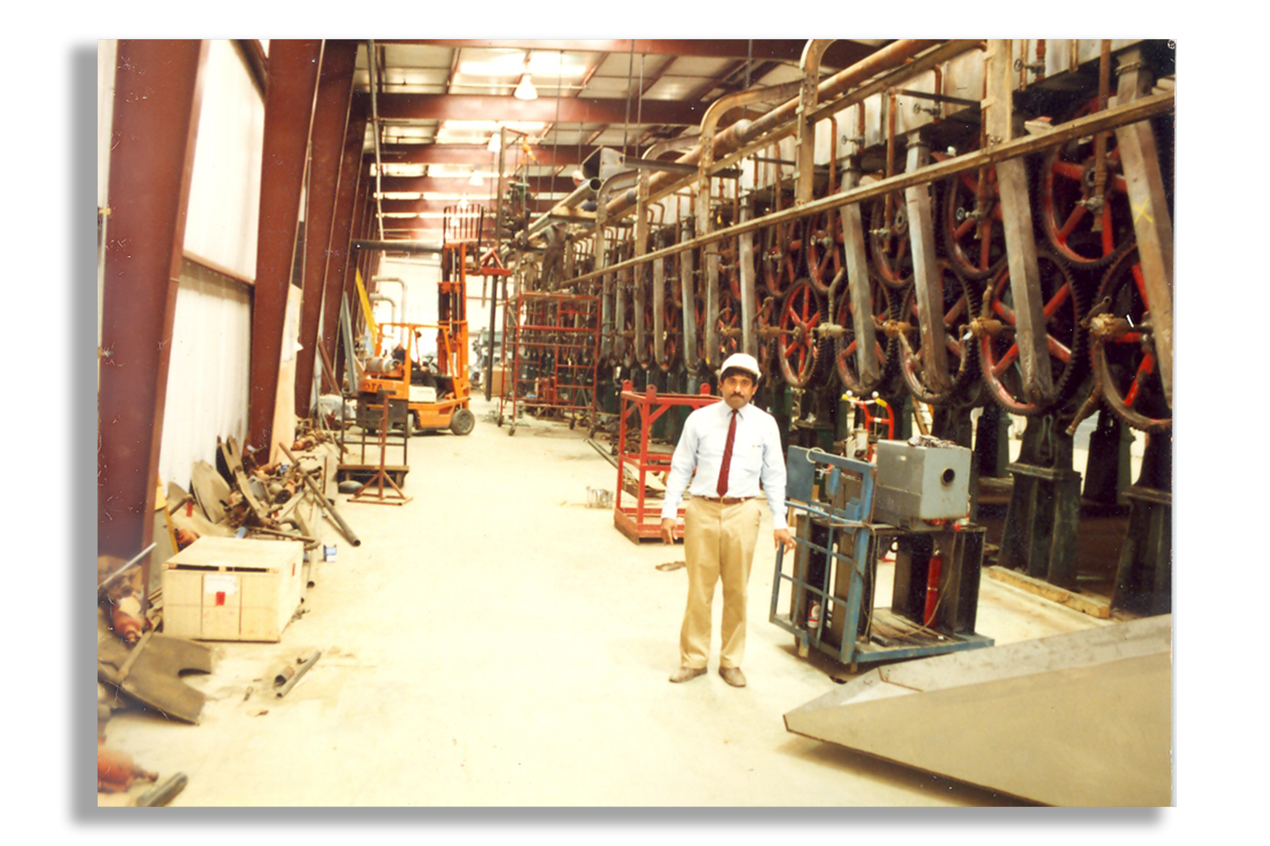 A man in business attire and a hard hat standing inside an industrial facility with large machinery, pipes, and equipment.